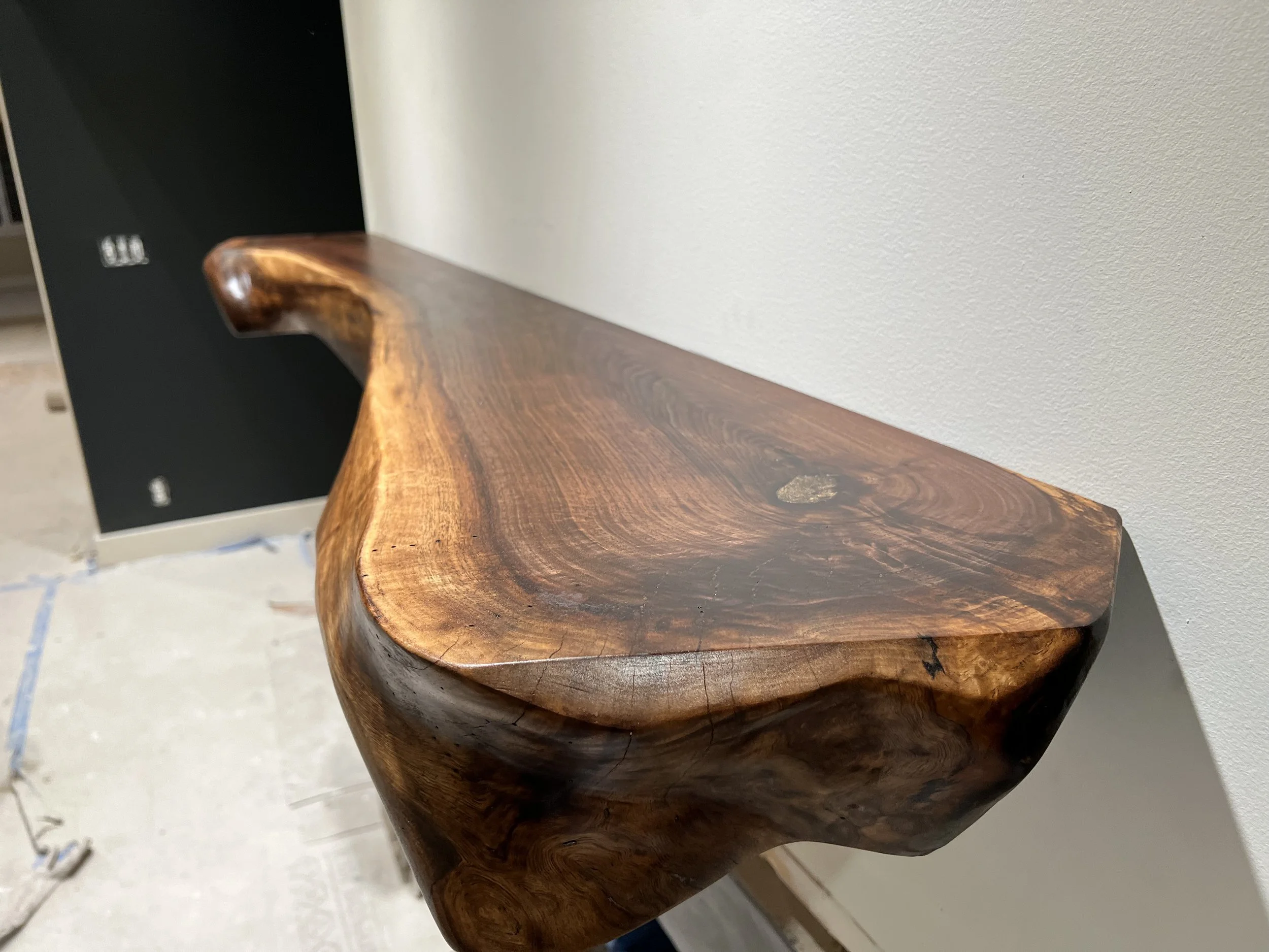 Close-up of a polished wooden shelf or table with natural curves and wood grain, leaning against a white wall.