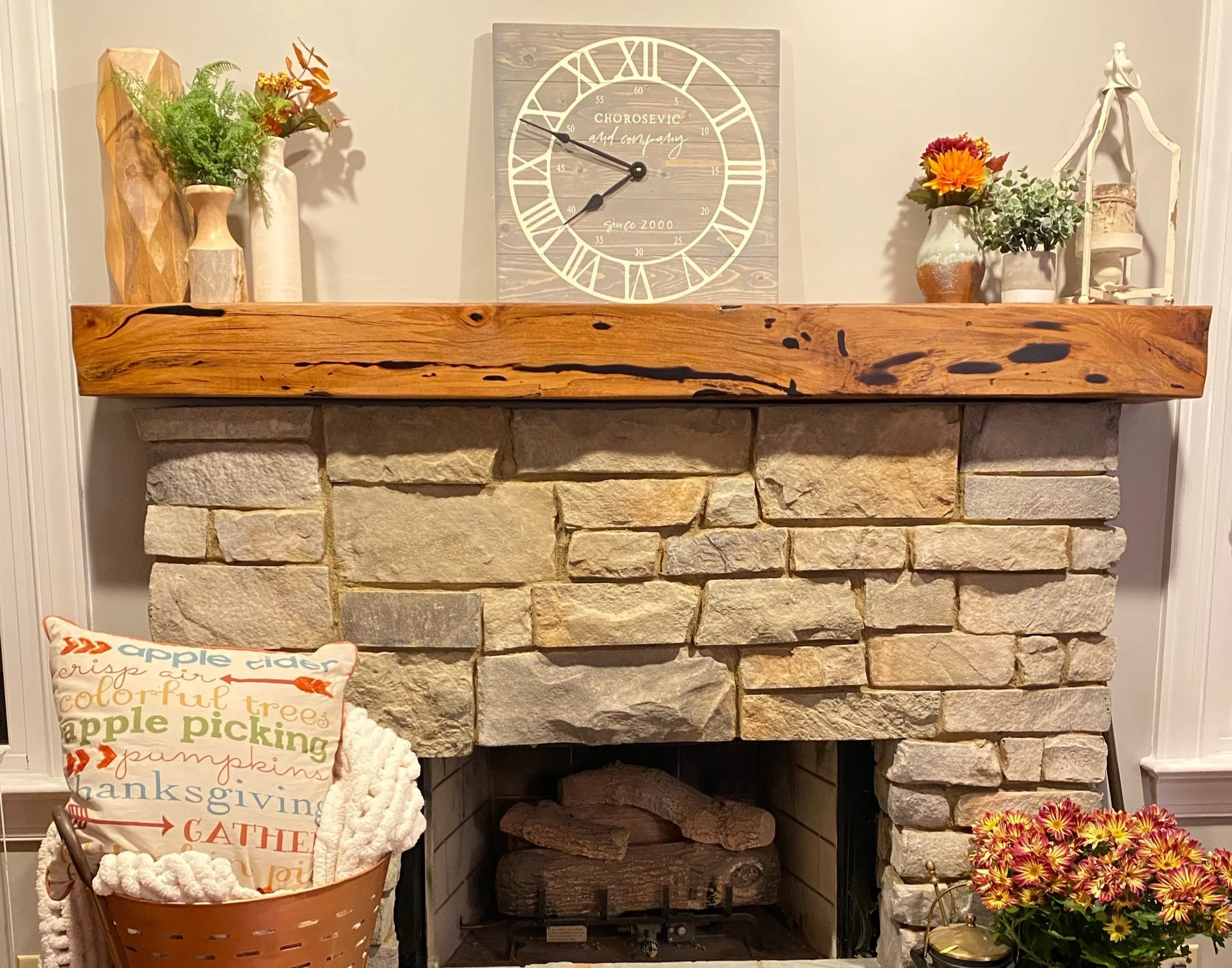 Decorative fireplace mantel with vases of flowers, a clock, and a basket of fall-themed pillows and blankets in front of a stone fireplace.
