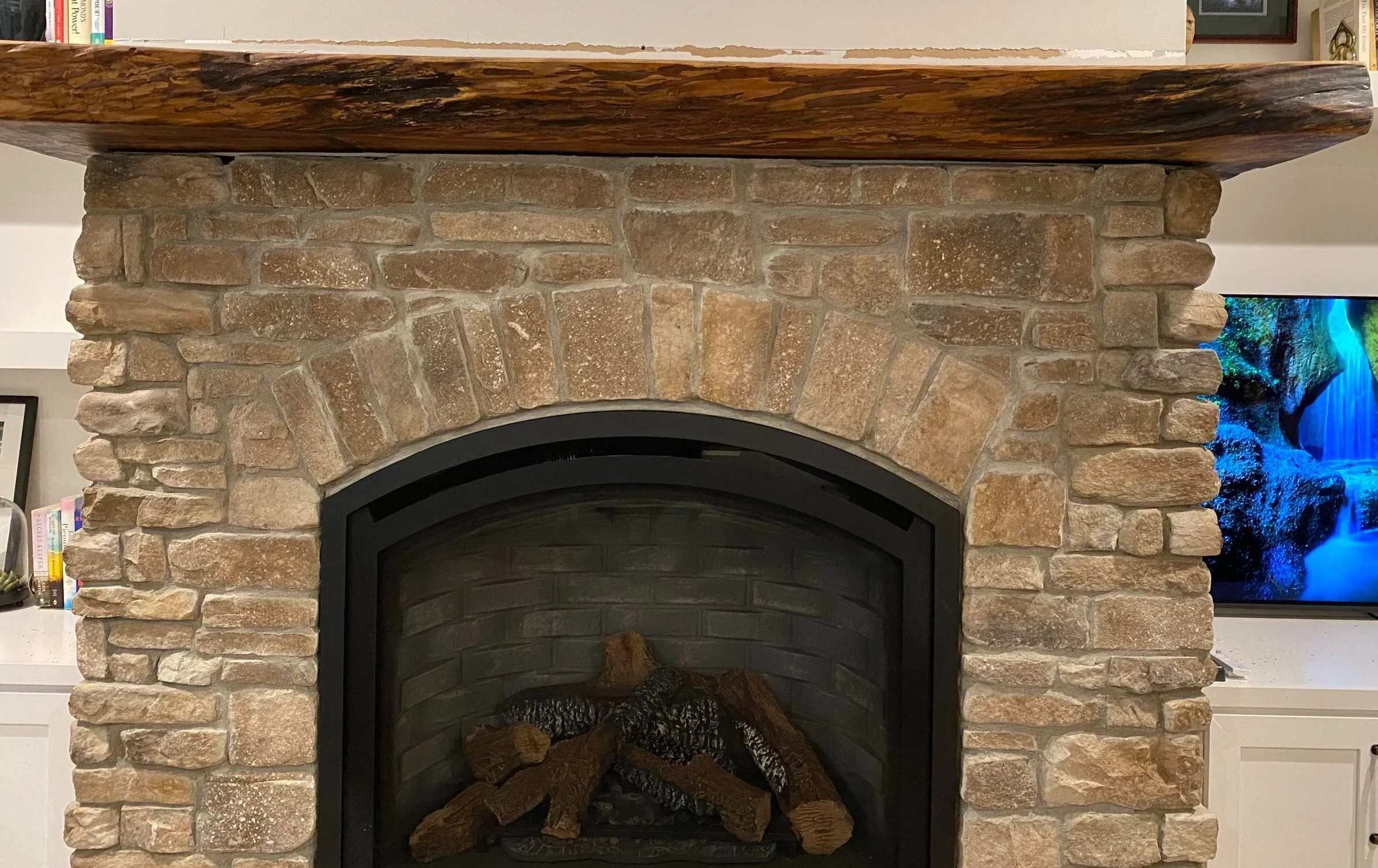 Close-up of a stone fireplace with a wooden mantel, with a television showing a waterfall scene to the right.