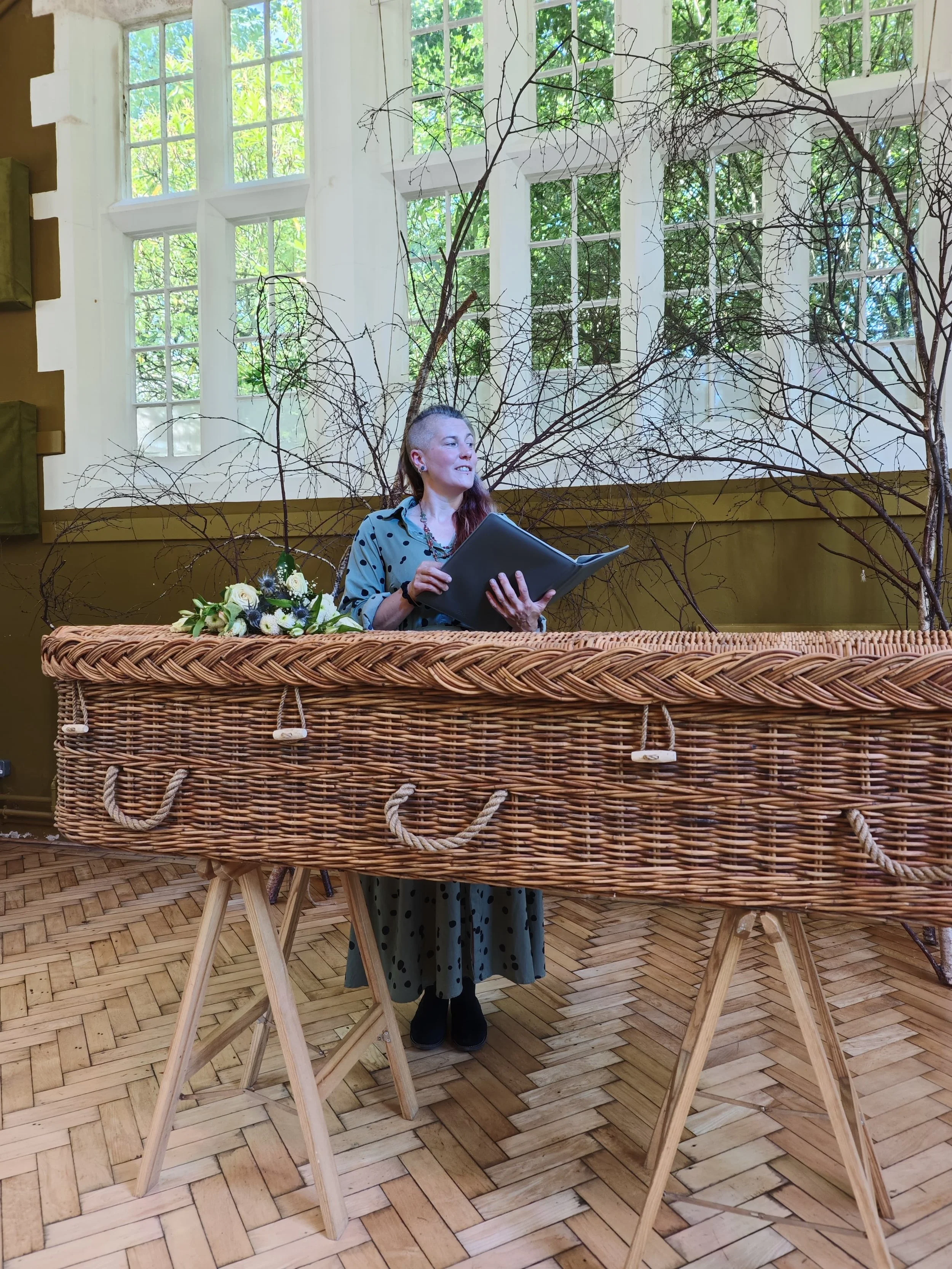 Emily, a white woman with red hair is standing behind a woven coffin in a large hall with some dried tree branches arranged behind her