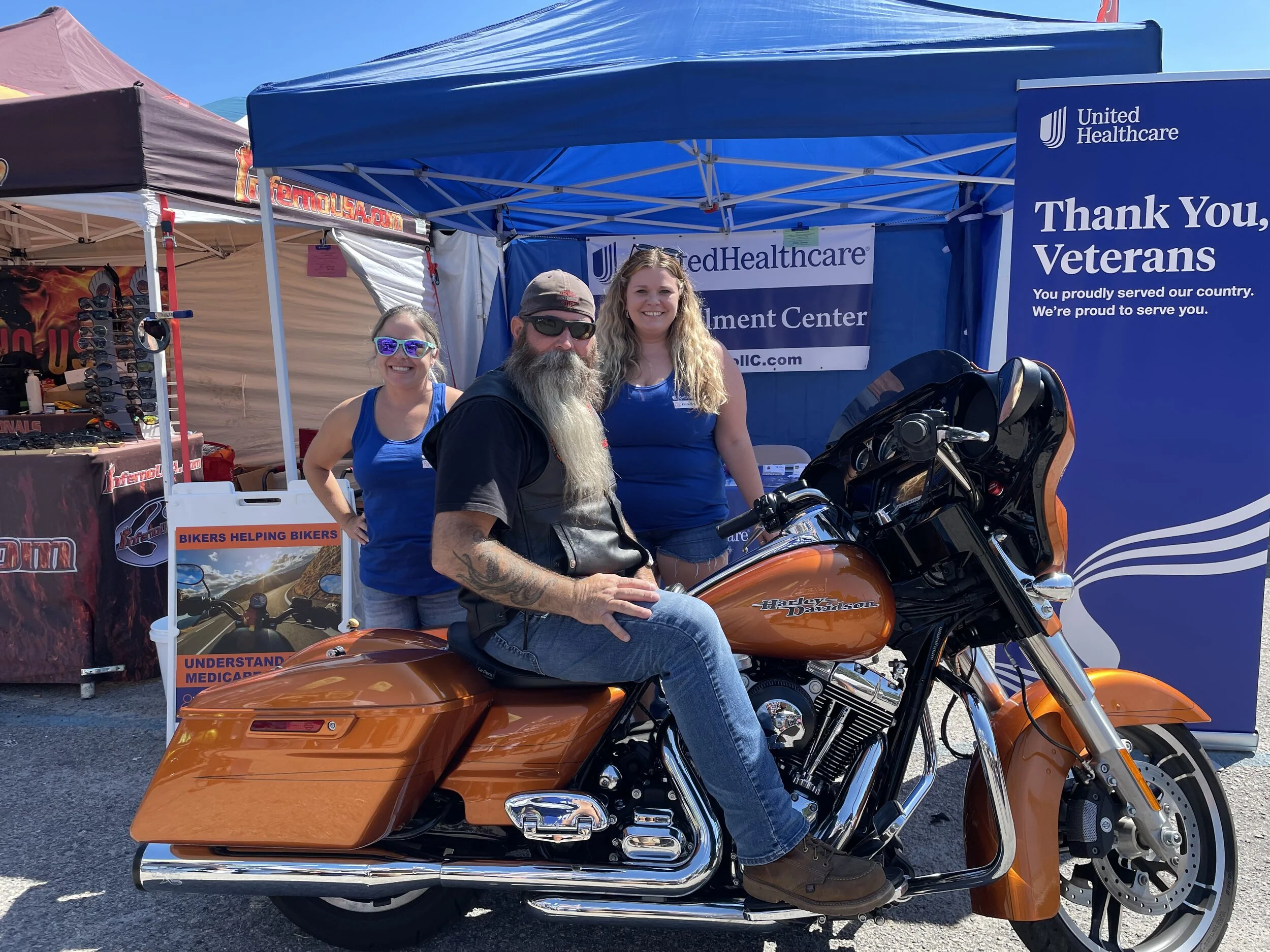 Three people standing behind a Harley Davidson motorcycle at a Veterans event. The person in front is sitting on the bike, wearing sunglasses, a black vest, and jeans, with long hair and a beard. Two women are standing behind him, wearing blue tank t