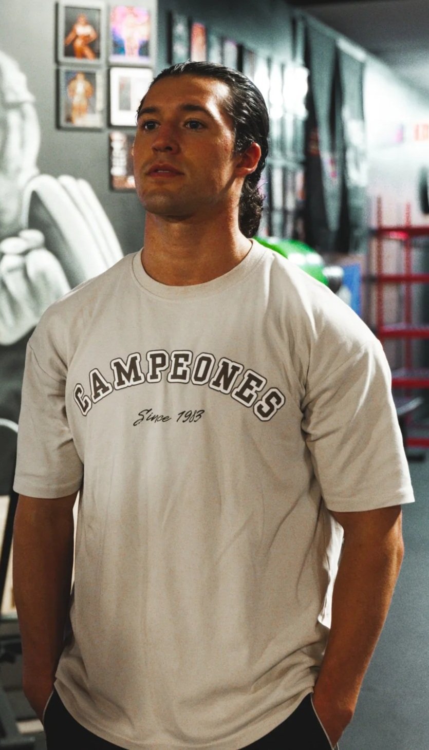 A man with dark, slicked-back hair wearing a white t-shirt with the word 'Campeones' and 'Since 1963' printed on it, standing inside a gym with framed pictures on the wall.