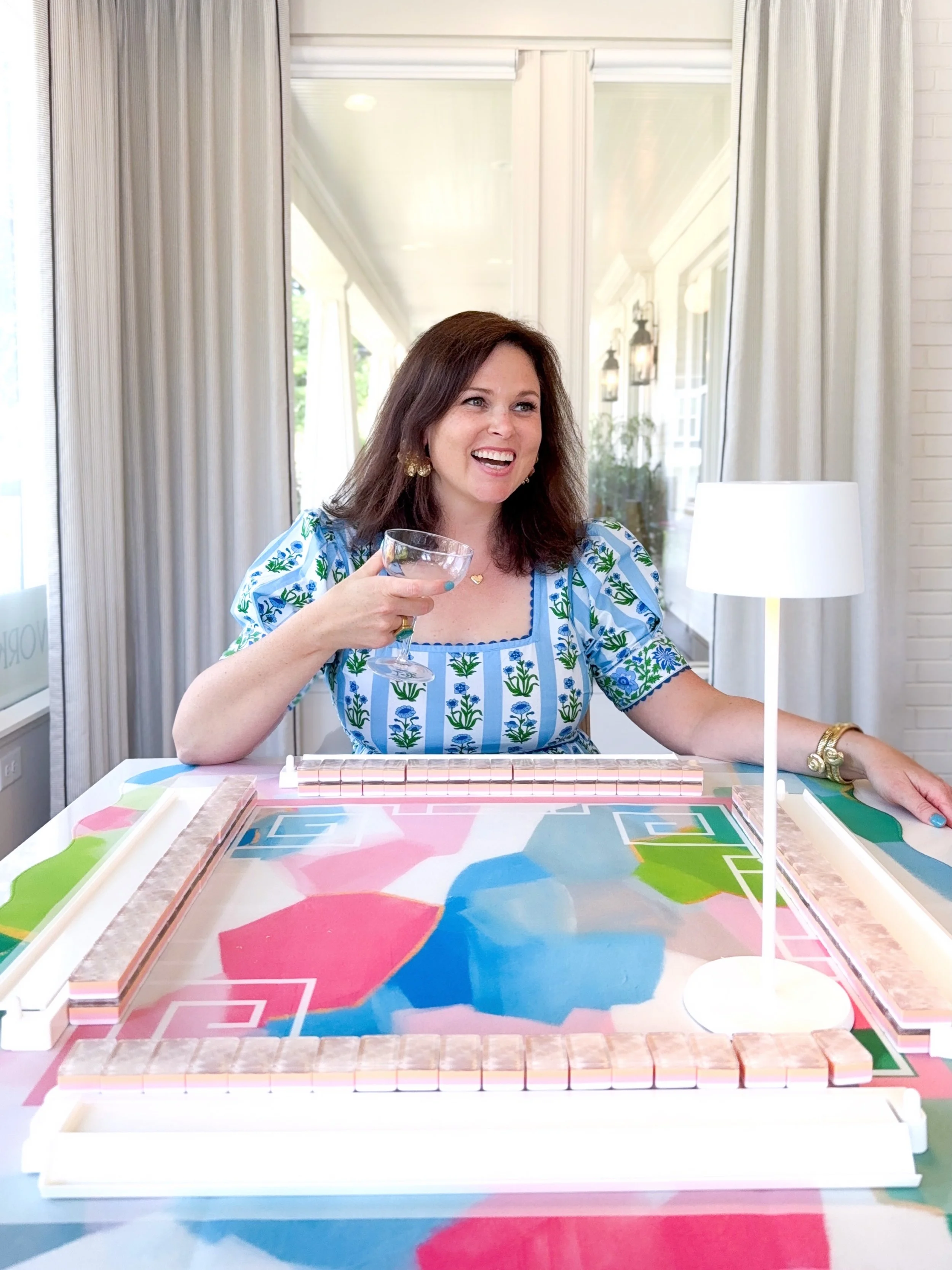 Woman smiling and raising a glass while playing a colorful board game on a table indoors with white curtains and a mirror in the background.