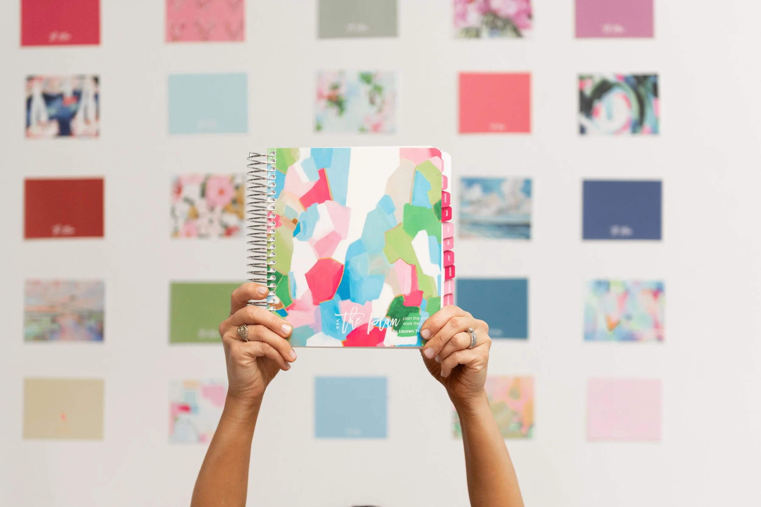 Person holding a colorful spiral-bound notebook with an abstract paint splatter design, against a backdrop of a wall with various colorful art prints.