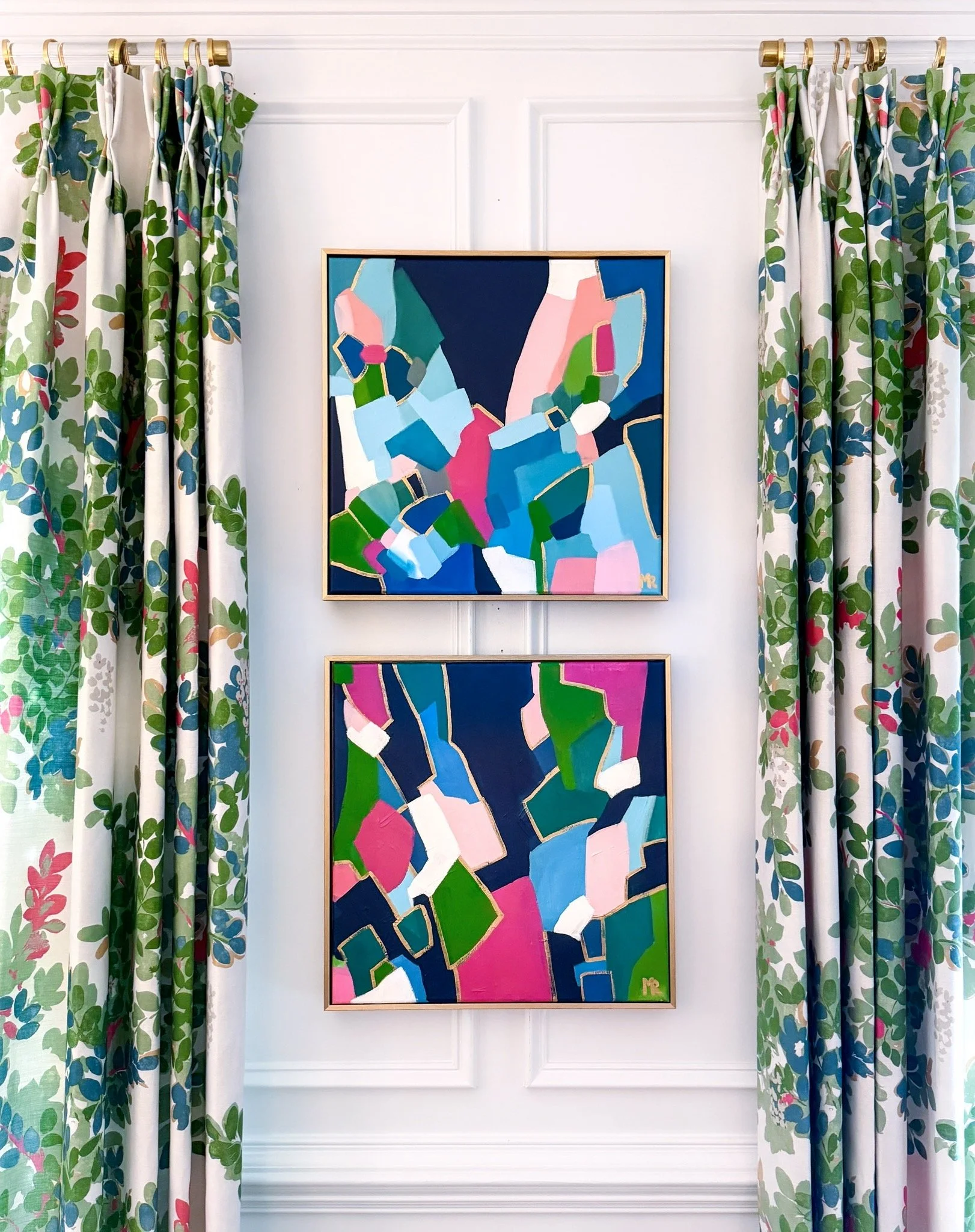 Colorful abstract paintings hanging on a white wall, framed in light wood, with floral patterned curtains on either side.