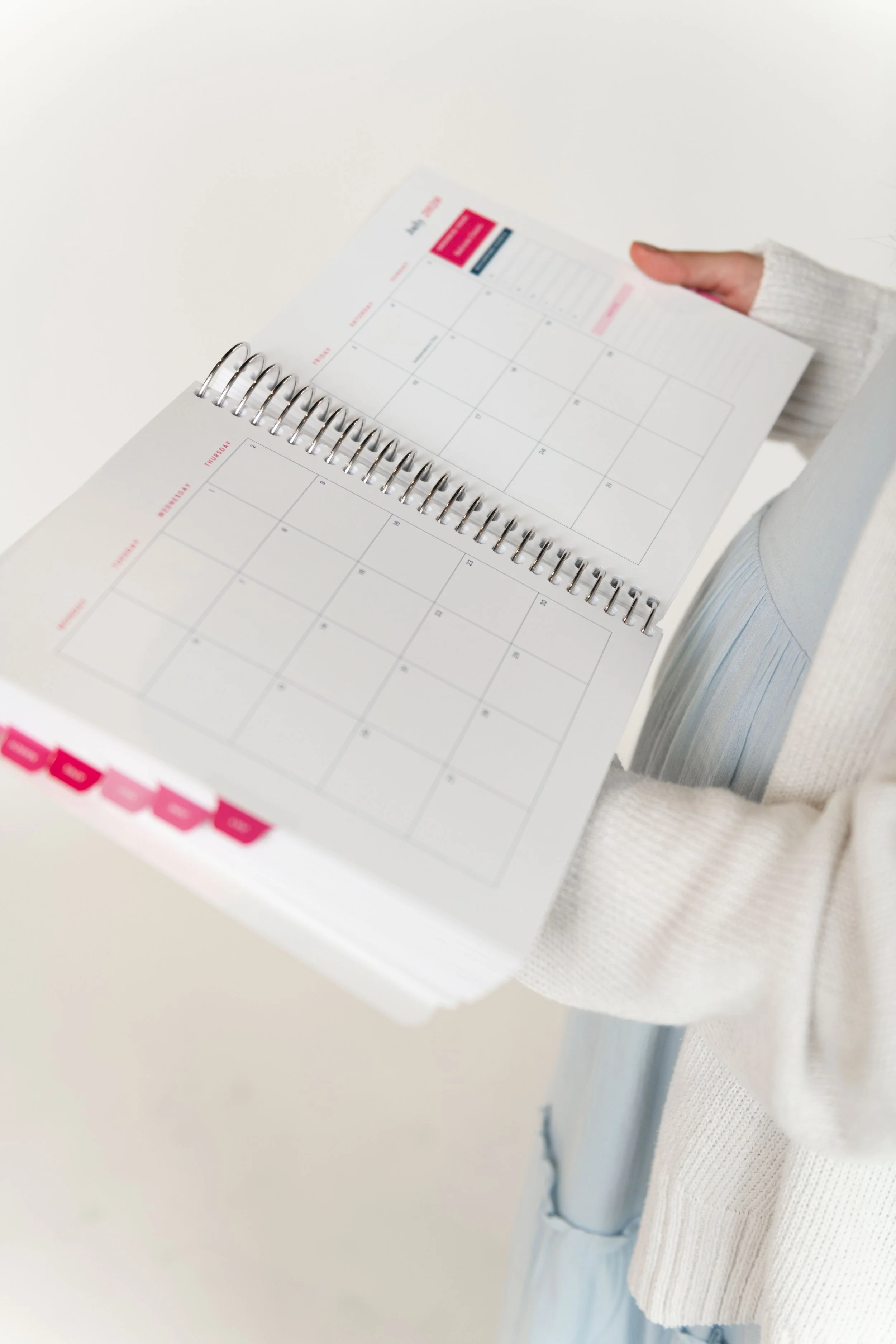 A person wearing a white sweater holding an open spiral-bound monthly planner with a blank calendar and a pink header on a white surface.