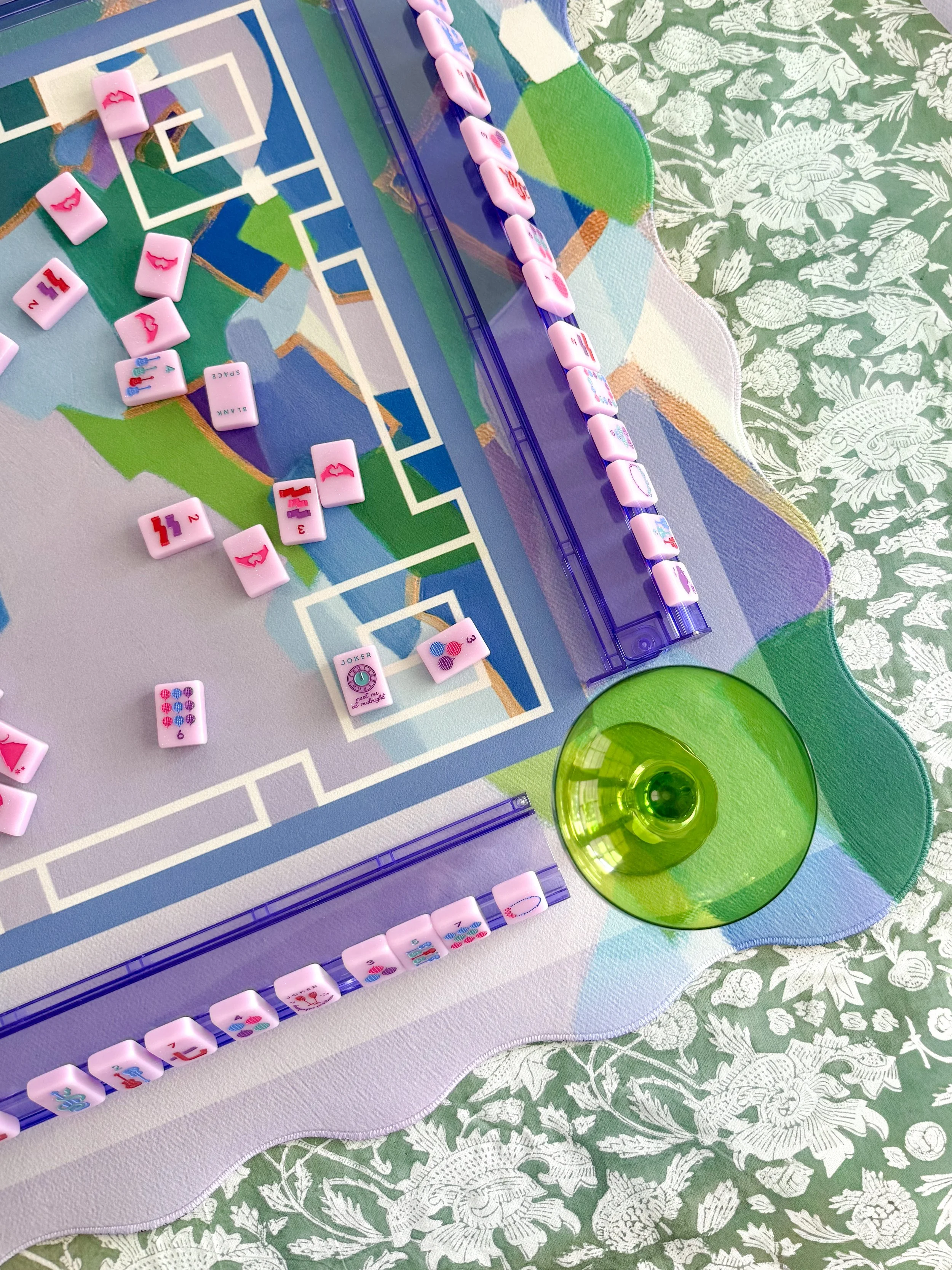 Close-up of a colorful tabletop game with pink domino-like tiles arranged on a board depicting a cityscape and some green and clear glass cups on a floral tablecloth.