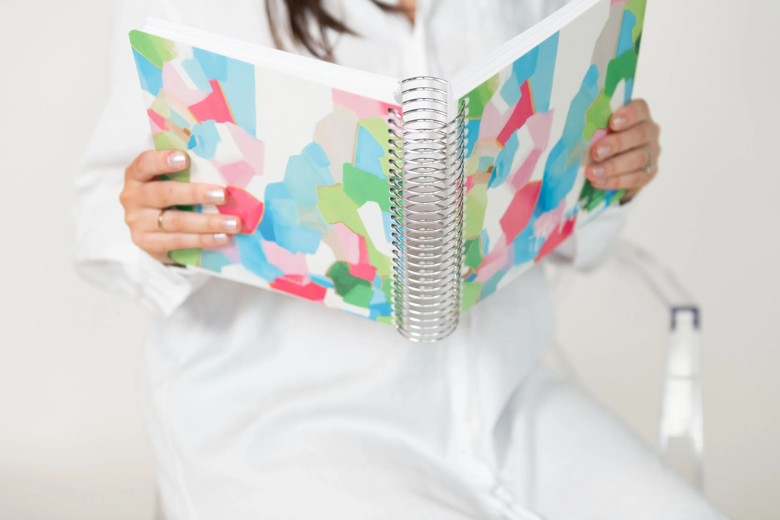 Person holding an open spiral-bound notebook with colorful abstract art on the cover.