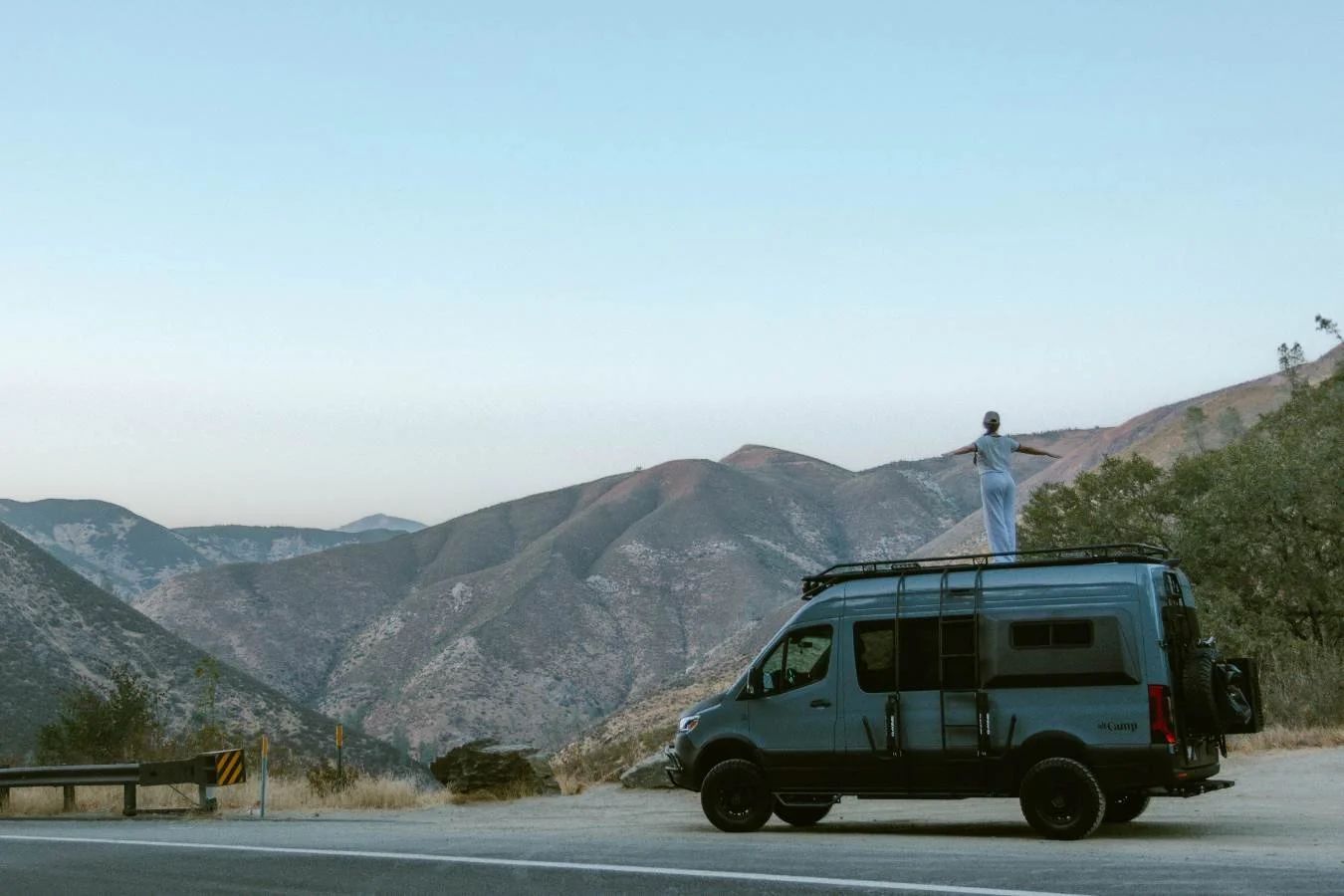 Sprinter van parked along mountain highway overlook with renter standing on roof. 
