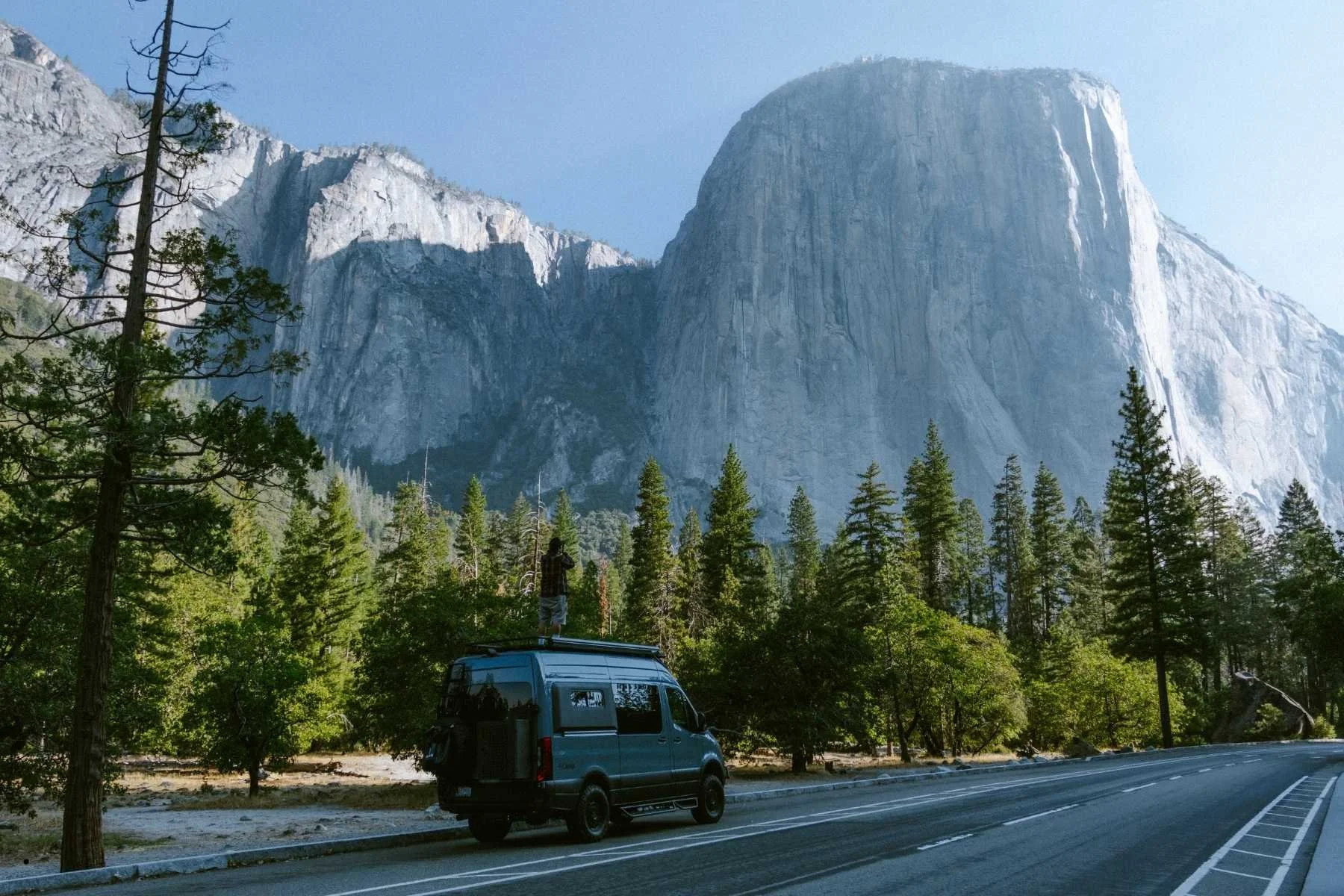 A person standing on the roof of a Sprinter van rental parked along a road in Yosemite National Park, California, with El Capitan's massive granite face rising above a pine forest in the background.
