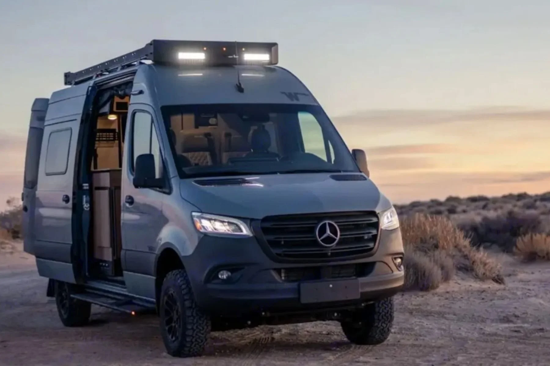 Luxury Mercedes Sprinter camper van rental in Las Vegas: A grey 2025 Winnebago Revel 4x4 parked in a desert landscape at sunset with lights illuminated.
