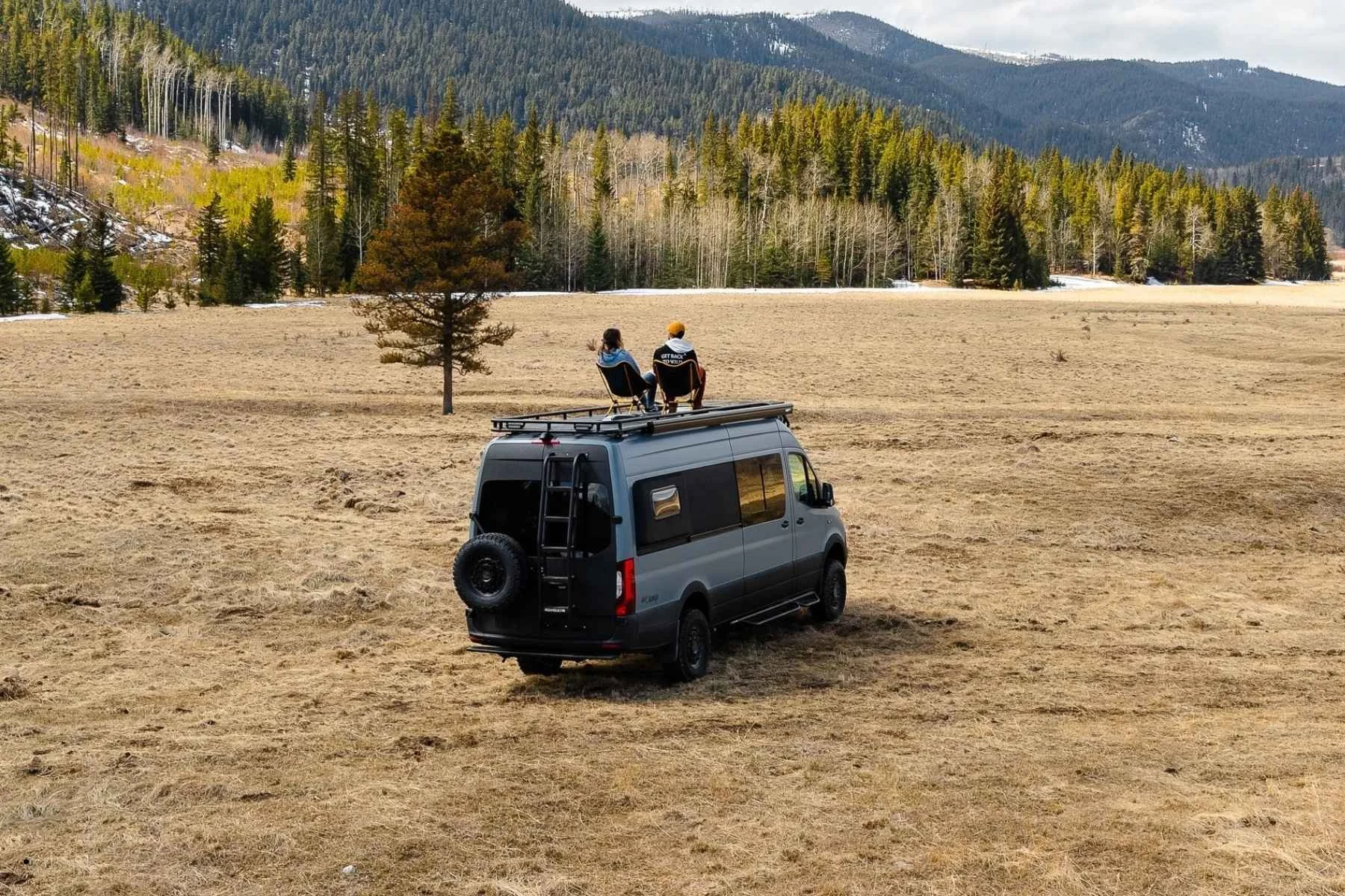 Two people relaxing on top of a Sprinter camper van rental parked in a Colorado mountain meadow.