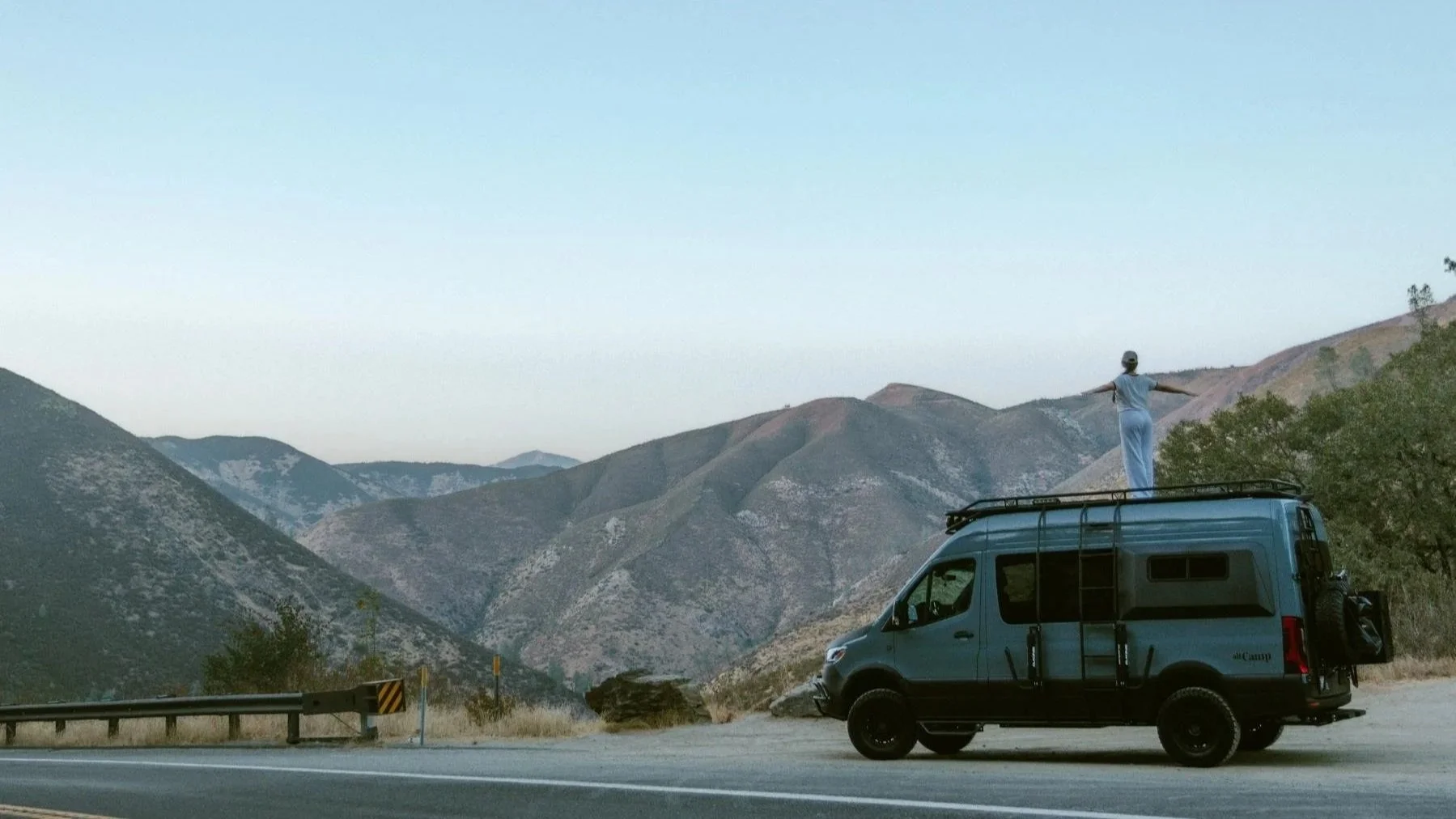 A blue Mercedes-Benz Sprinter camper van parked on a mountain roadside at dusk, with a person standing on the roof rack with arms outstretched, looking out over rolling mountain ranges in the distance..