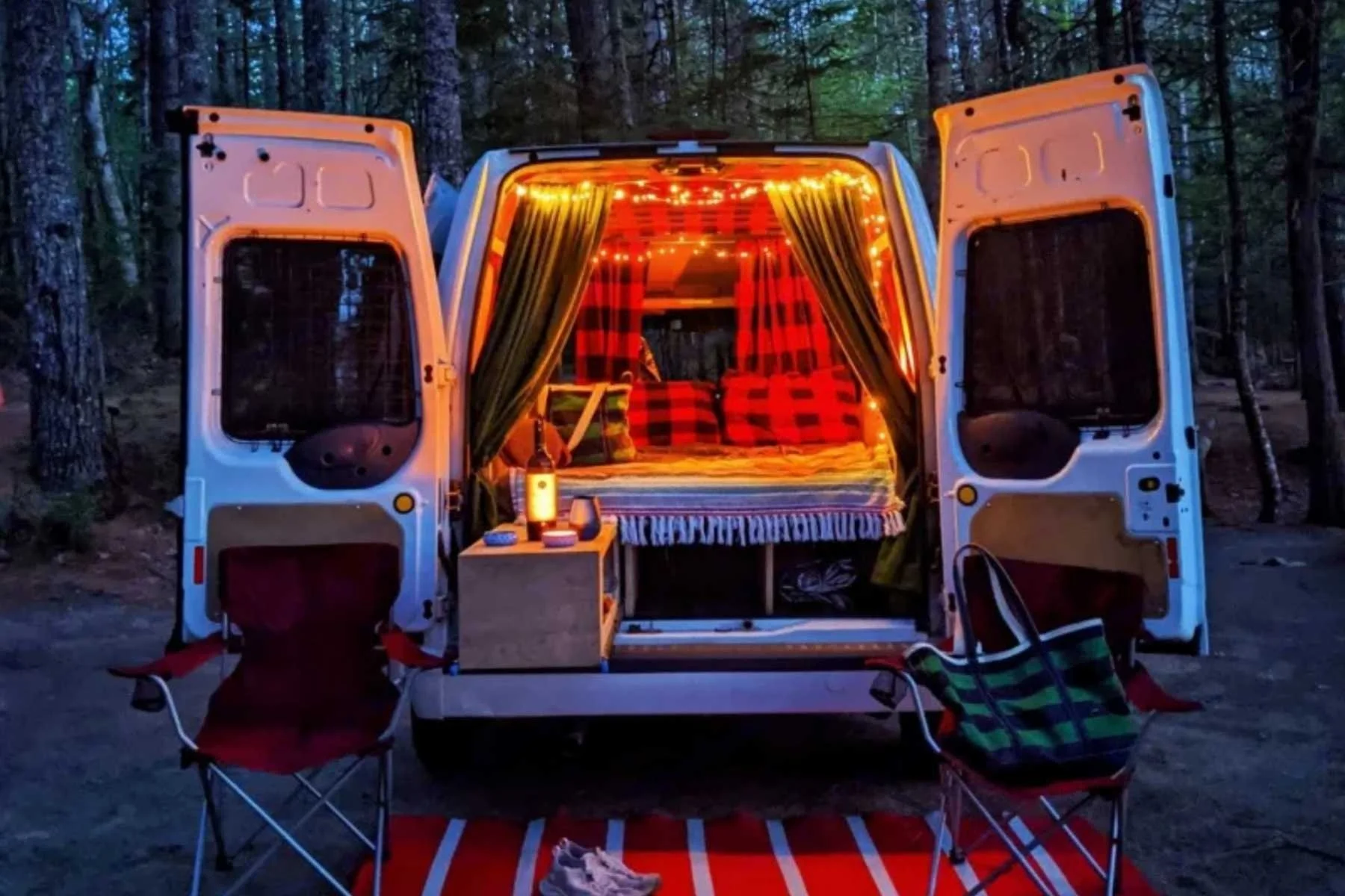 Best small conversion van in Maine set up at a forest campsite with a side awning, outdoor chairs, and a table outside the sliding door.