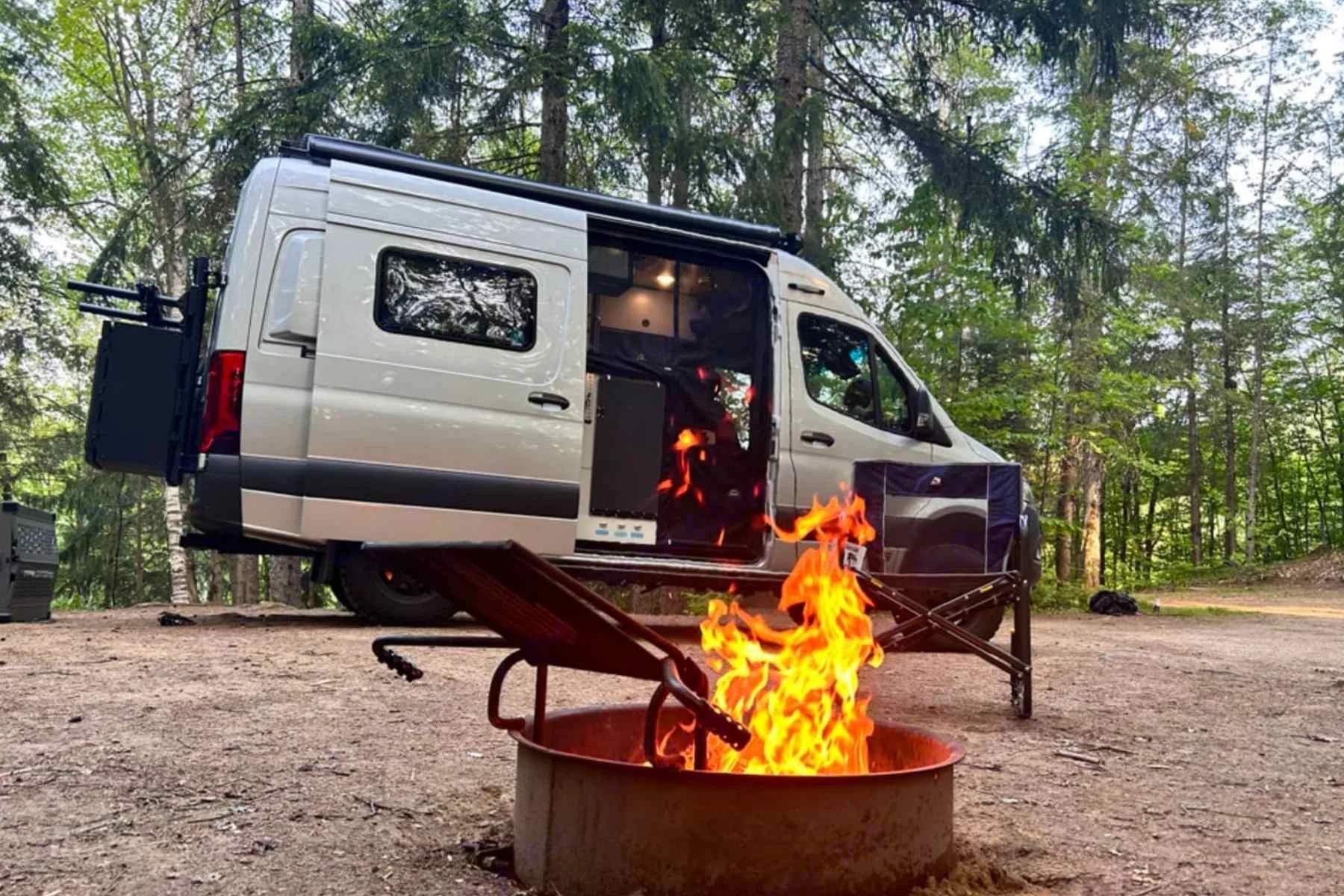 Sprinter rental in Maine parked at a forest campsite with the side door open and a campfire burning in the foreground.