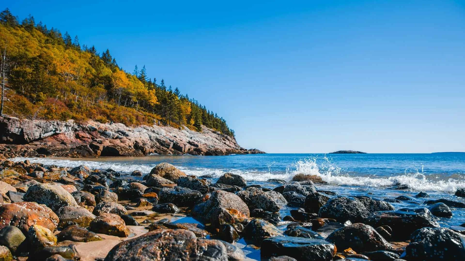 Rocky Maine coastline with ocean waves, forested cliffs, and clear blue sky near a scenic beach destination.