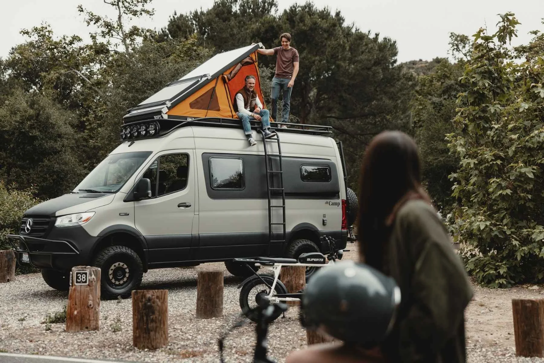 Two people sitting on top of a Mercedes-Benz Sprinter camper van with a rooftop tent open at a campsite, with an altCamp-branded van conversion, ladder, roof rack, and a Super73 electric bike parked alongside.