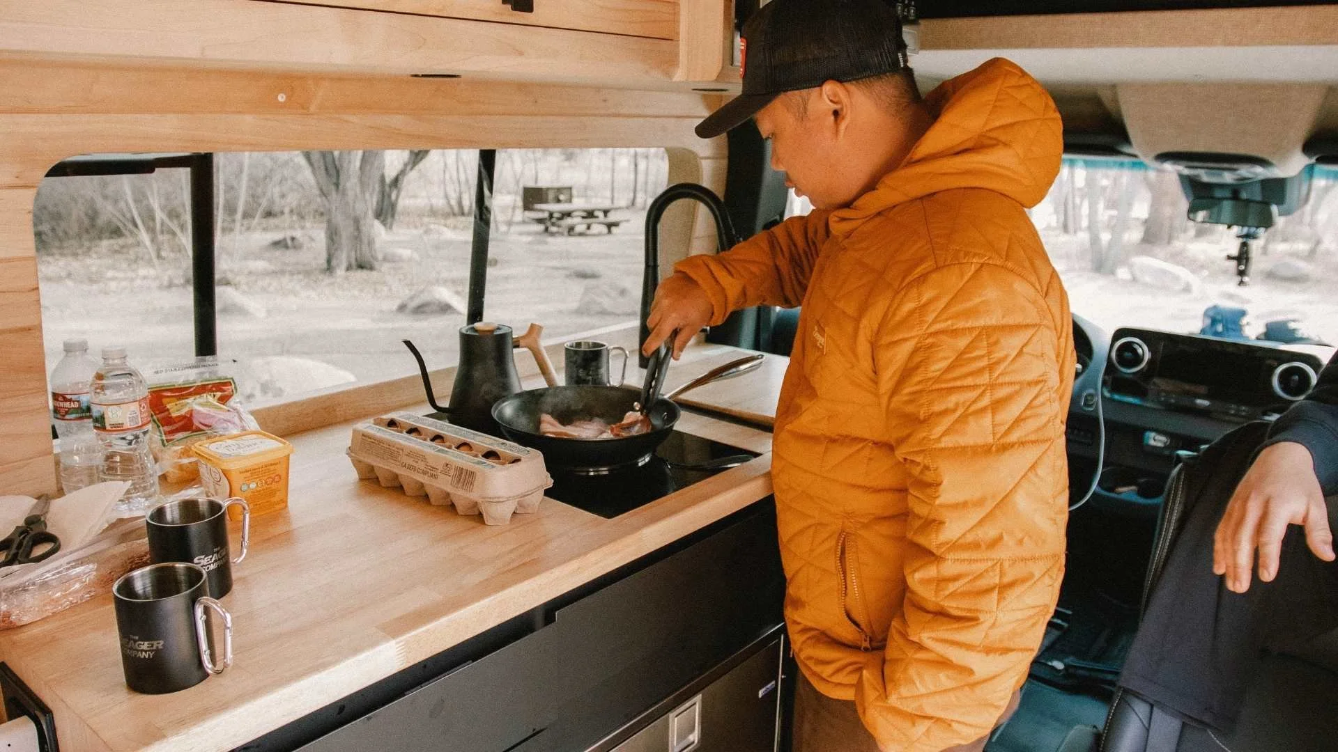 A man wearing an orange jacket and black cap cooks bacon in a skillet inside a camper van kitchen. The countertop holds eggs, butter, and coffee mugs, with a wooded campsite visible through the window.