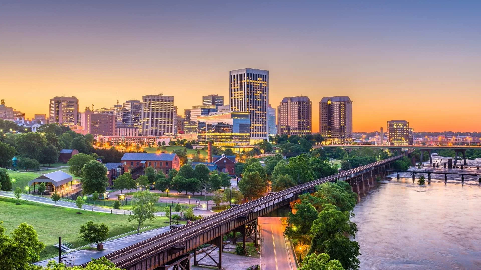 Richmond, Virginia skyline at sunset with modern downtown buildings overlooking the James River and historic railroad bridge in the foreground.