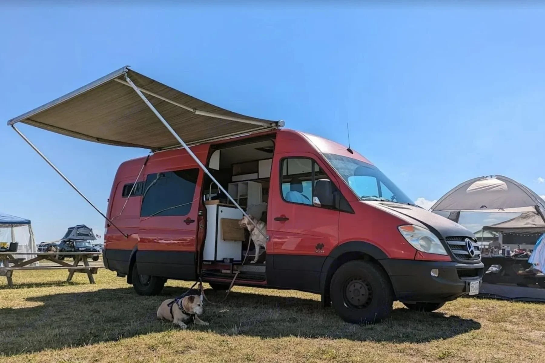 Top-rated luxury Sprinter van conversion rental in Virginia with side awning extended at a sunny campsite, featuring an open sliding door and cozy interior setup for outdoor adventures.