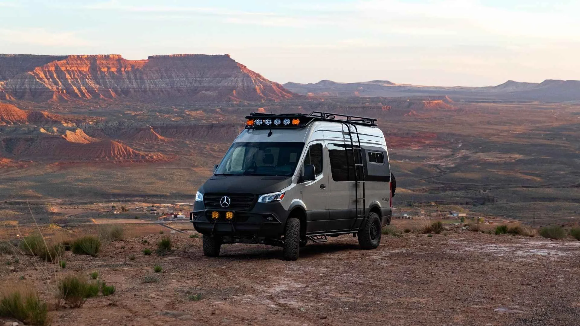 A silver Mercedes Sprinter camper van rental parked on a cliffside overlooking the rugged desert plateaus and red rock canyons of Utah at sunset.