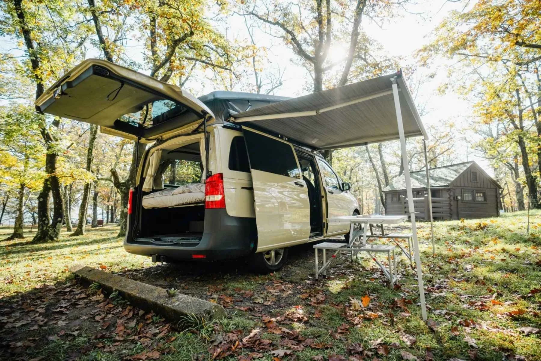 Best-rated budget Mercedes Metris camper van rental in Virginia parked at a wooded campsite with rear hatch open, side awning extended, and outdoor table set up for a cozy forest camping experience.