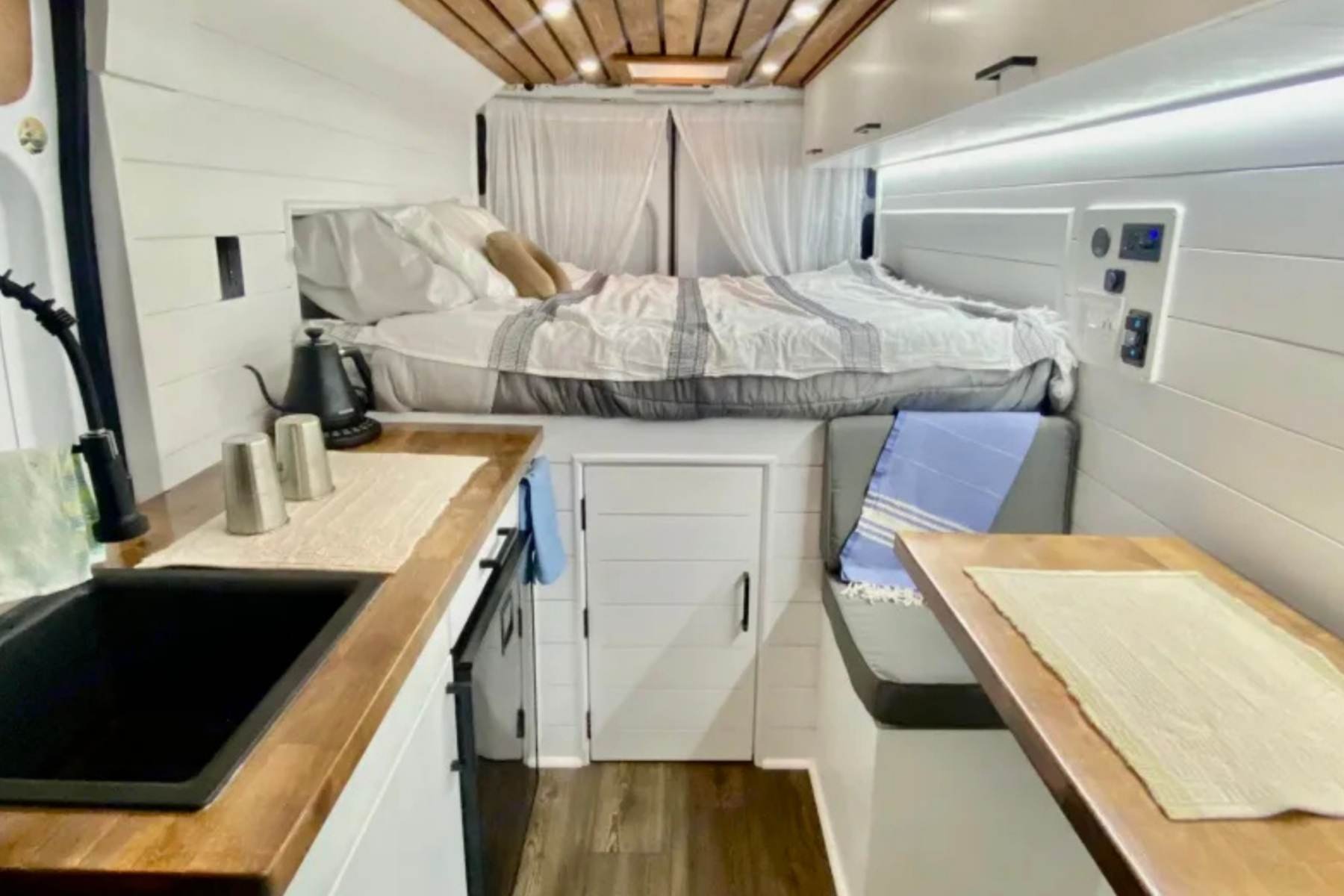 Interior of the Vandal camper van with cozy bed, wood cabinets, kitchenette, and dining bench in a warm, homey layout.