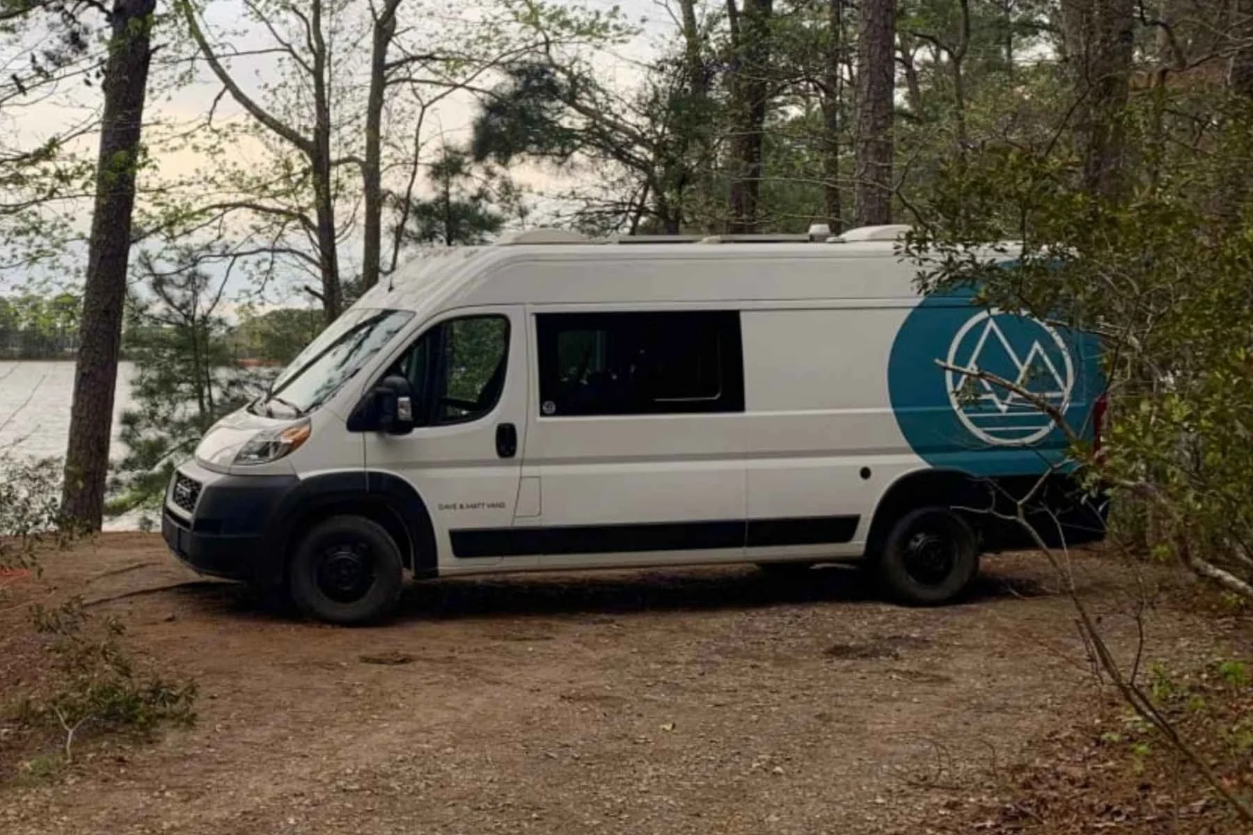 Top rated camper van rental in South Carolina parked at a lakeside campsite, featuring a white conversion van surrounded by tall pine trees and scenic waterfront views.