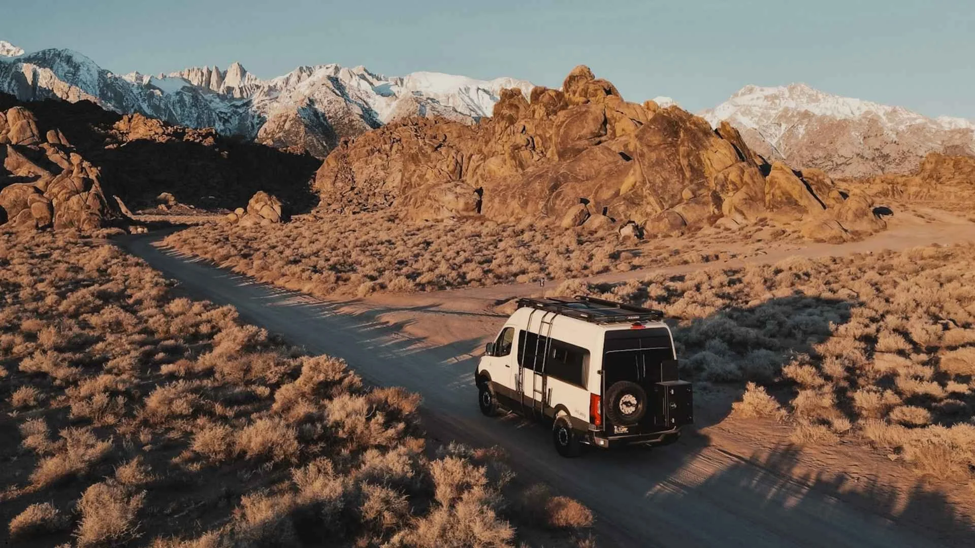 A white camper van driving a dirt road through the Alabama Hills with snow-capped Sierra Nevada mountains in the background