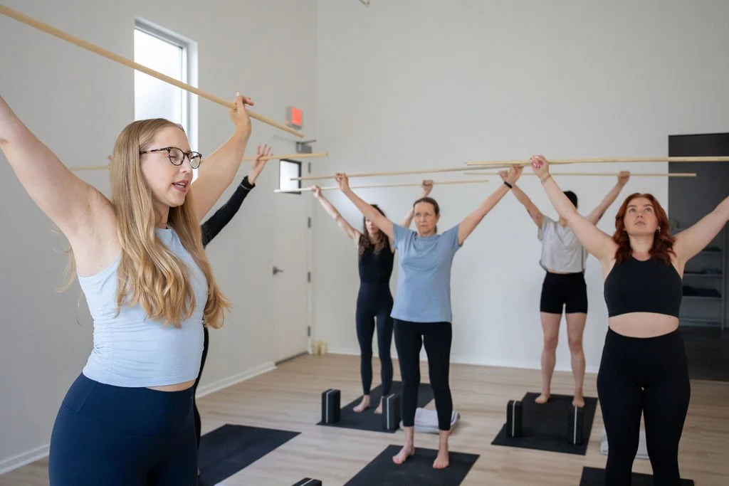A female teacher leads five students in mobility exercises using a wooden dowel.