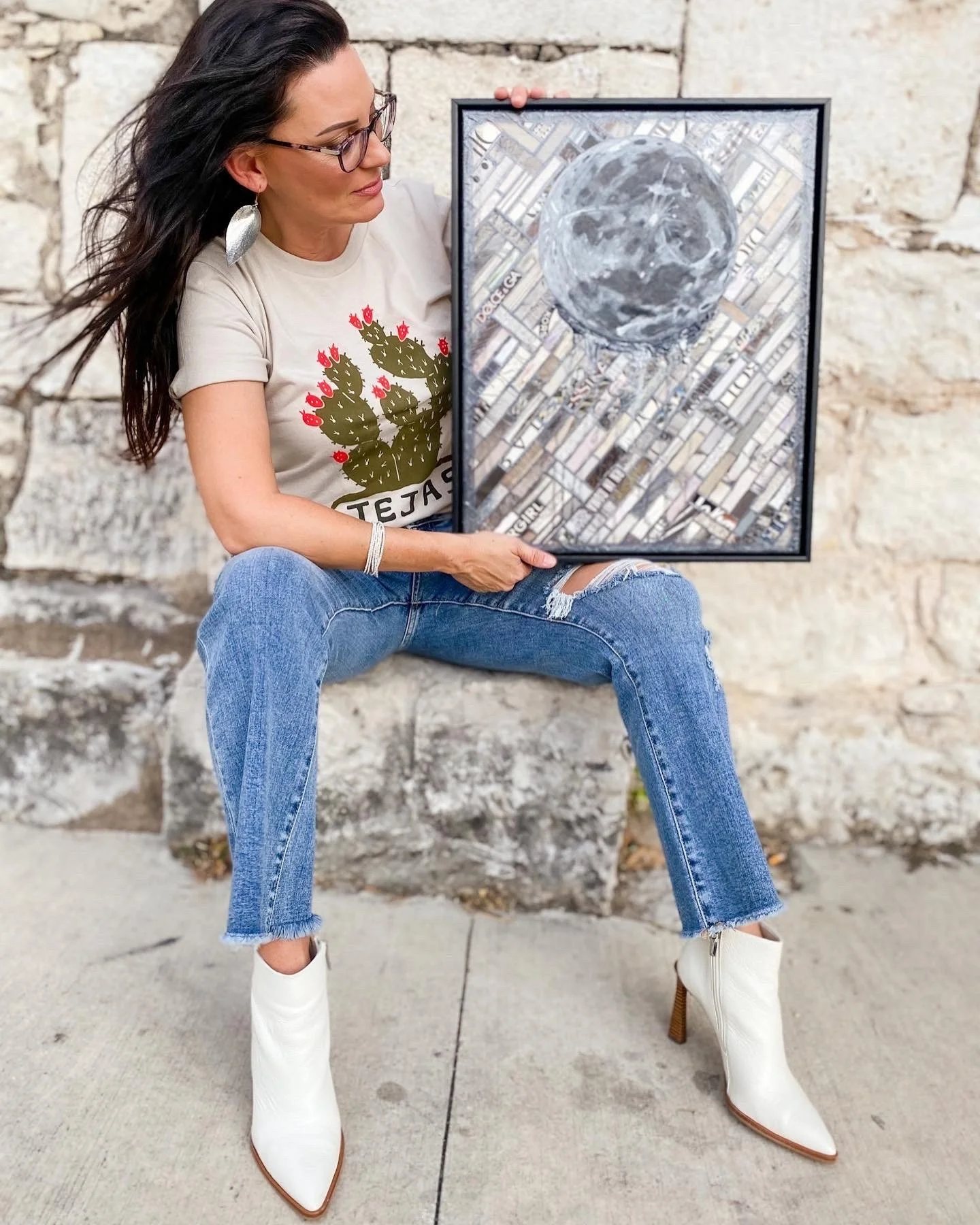 Woman sitting outdoors against a stone wall holding framed artwork of the moon and cityscape.