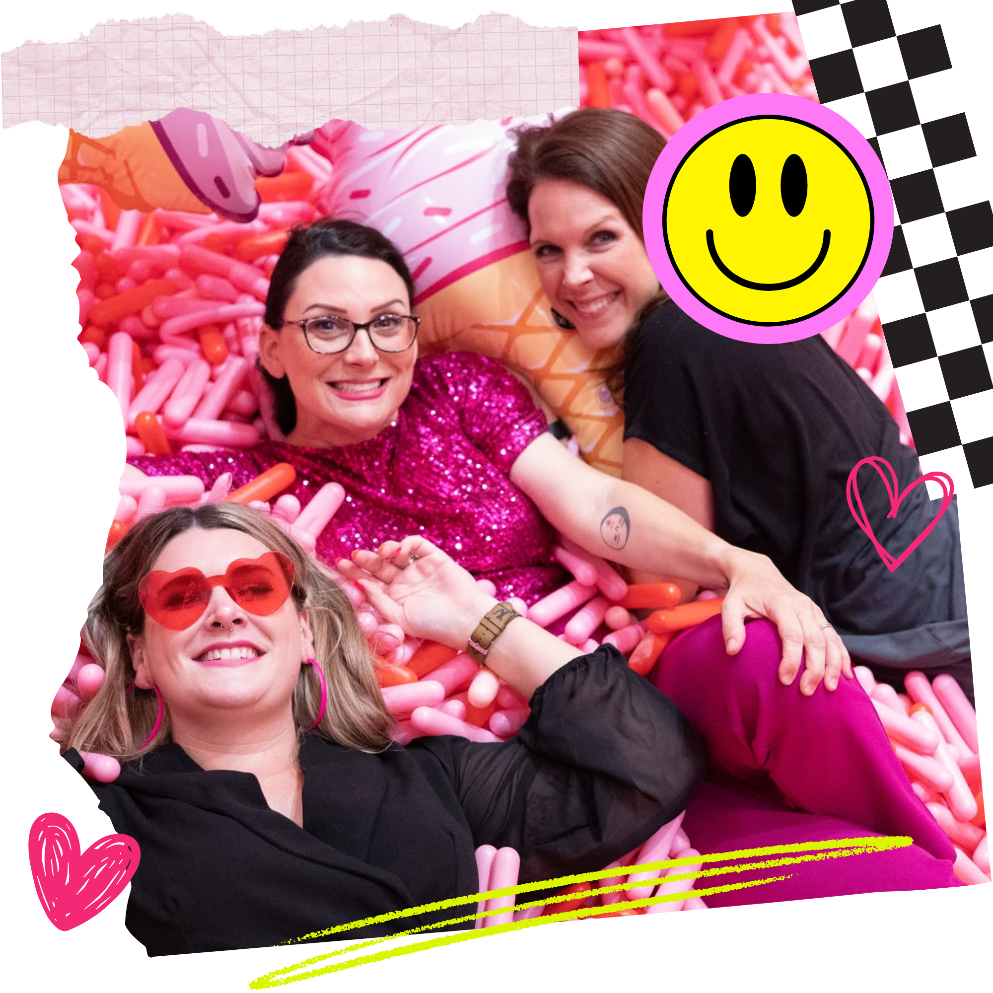 Three women smiling and lying on a bed of pink and orange balloons, with one wearing heart-shaped sunglasses and bright pink earrings, and another with glasses and a sequin top, all embracing each other cheerfully.