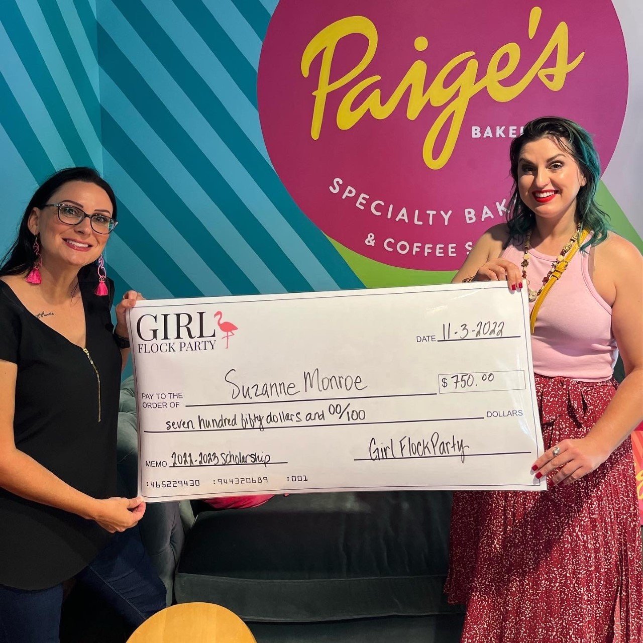 Two women standing indoors holding a large check made out to Suzanne Monroe for $750, dated November 3, 2022, for a Girl Flock Party scholarship. The background features a colorful sign for Paige's Bakery and Coffee Shop.