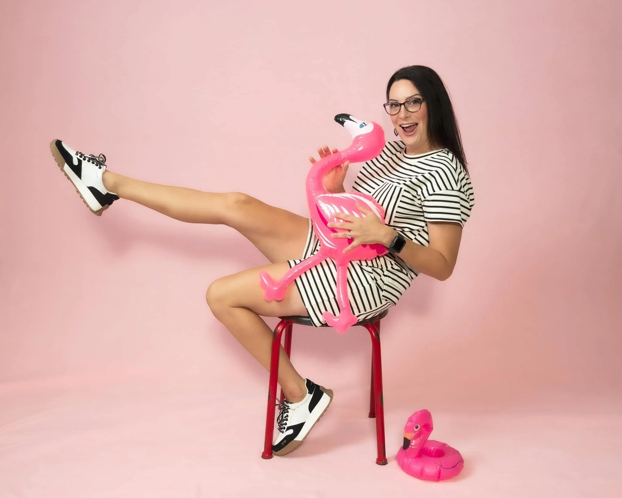 A woman with long dark hair wearing glasses and a striped shirt is sitting on a red stool against a pink background. She is holding a pink inflatable flamingo around her waist, with one leg raised and wearing black and white sneakers. There is a second pink inflatable flamingo on the floor in front of her.
