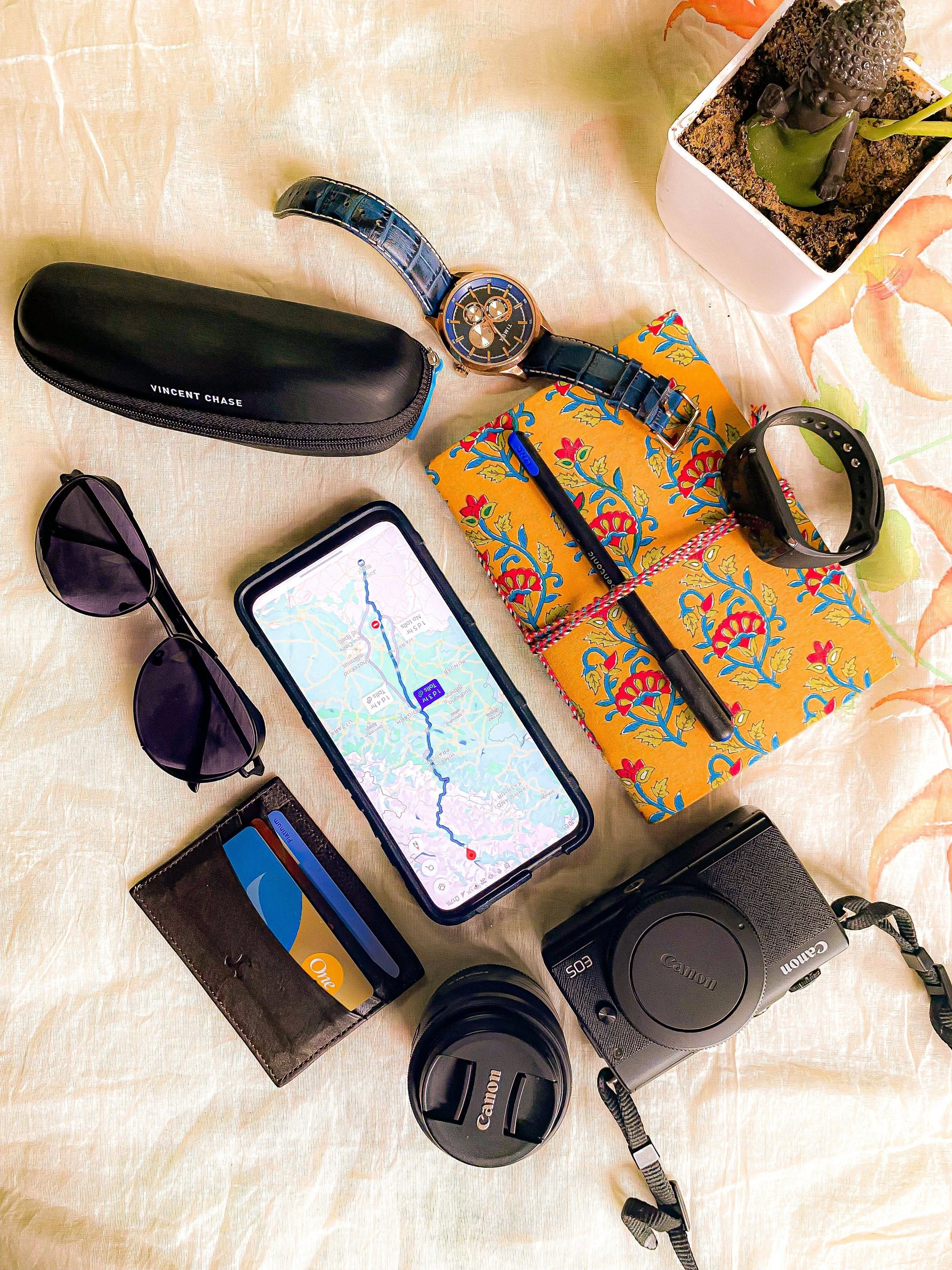 A flat lay of travel and photography items on a light-colored surface, including sunglasses, a smartphone displaying a map, a black case labeled 'VINCENT CHASE,' a wristwatch with a blue strap, a colorful patterned box with a pen and a black smartwatch on top, a camera with a lens cap, and a potted succulent plant in a white square pot.