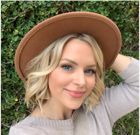 Blonde woman wearing a brown hat and beige sweater, smiling in front of green foliage.