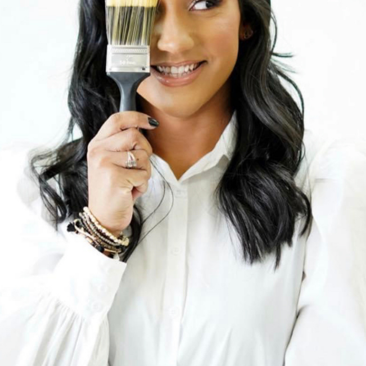 Woman smiling, holding a paintbrush in front of one eye, wearing a white blouse and bracelets.