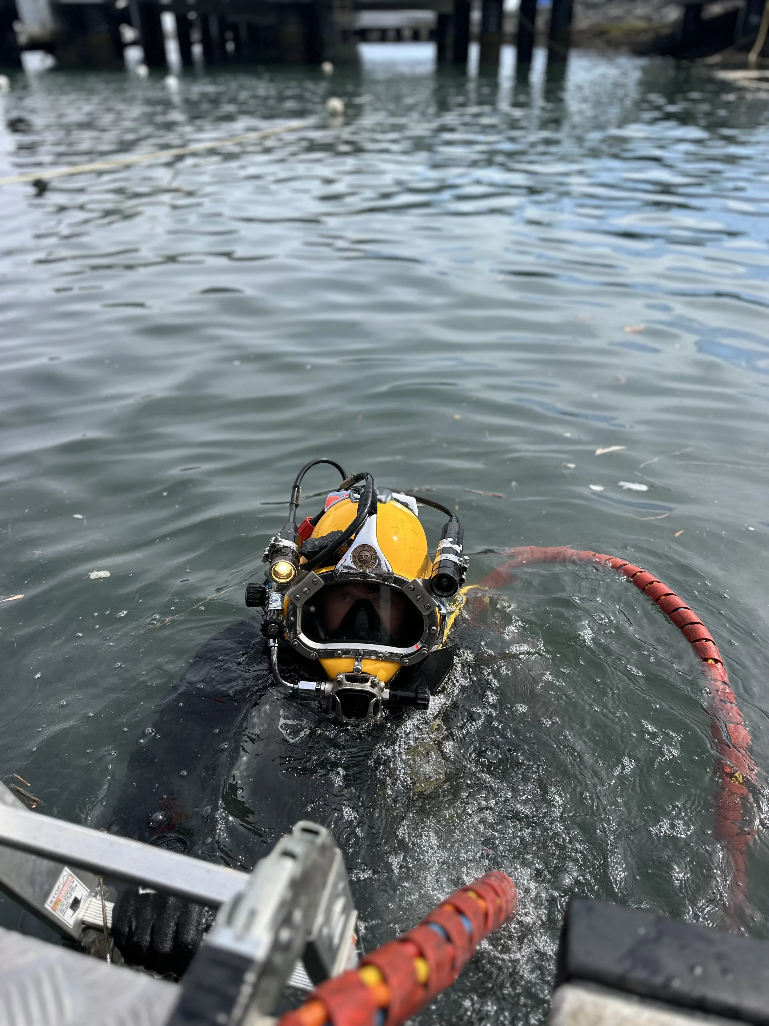 A diver in a yellow suit and breathing gear emerging from the water near a dock or pier in a harbor or marina.
