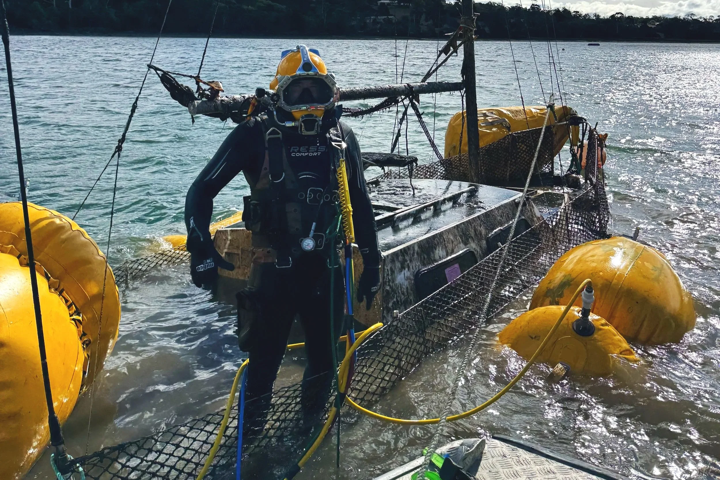 A diver wearing a wetsuit and helmet standing next to a sunken yellow submarine with large yellow flotation devices on the water.