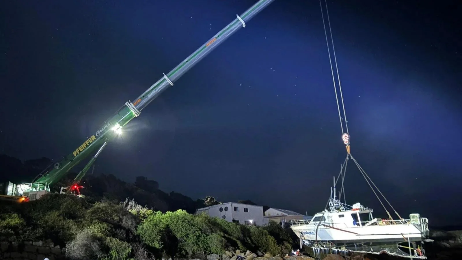 A large green crane lifting a yacht at night, with a house and trees in the background.