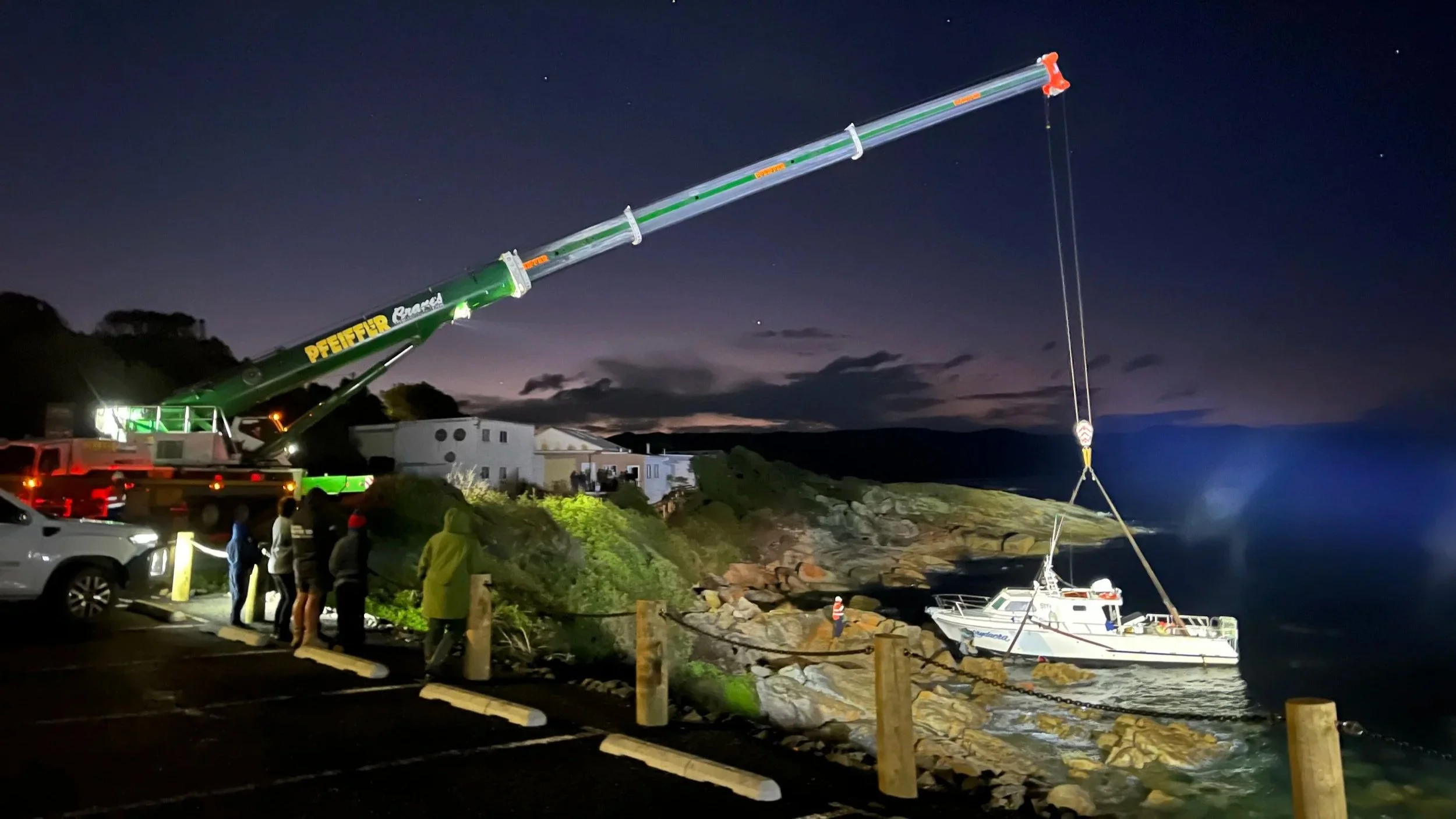 A boat is being lowered into the water by a crane at night near a rocky shoreline, with several onlookers watching.