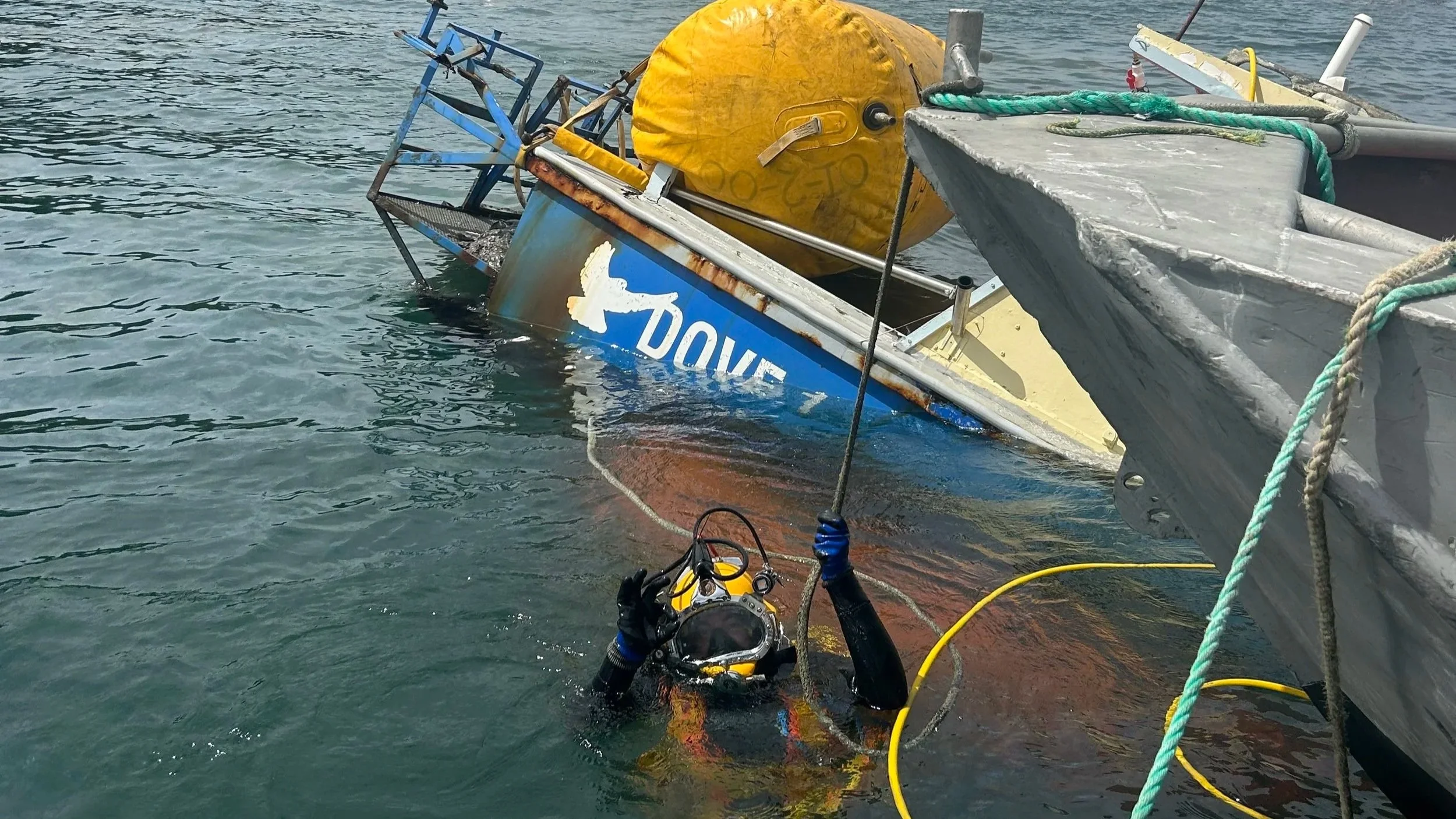 Person in diving gear underwater near a boat, holding an underwater camera or equipment, with a partially submerged yellow buoy and some additional boat parts visible.