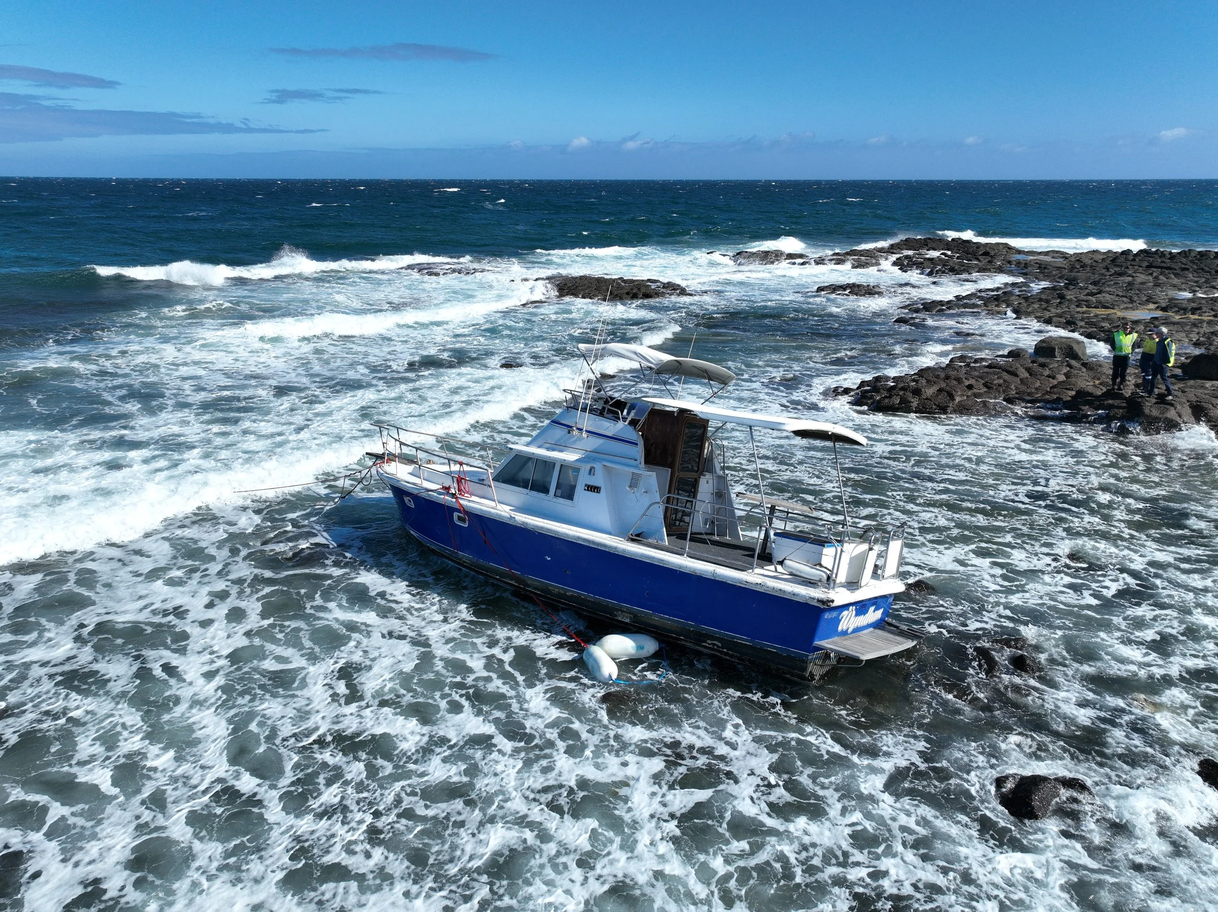 A blue and white boat is stranded on a rocky shoreline with waves crashing around it. Three people are standing on the rocks nearby, inspecting the area.