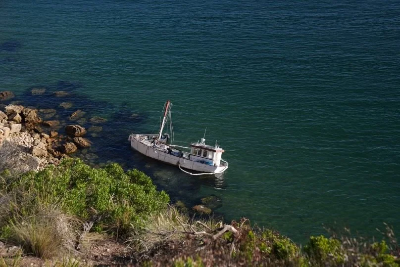 A boat anchored near a rocky and green shoreline in clear blue water.