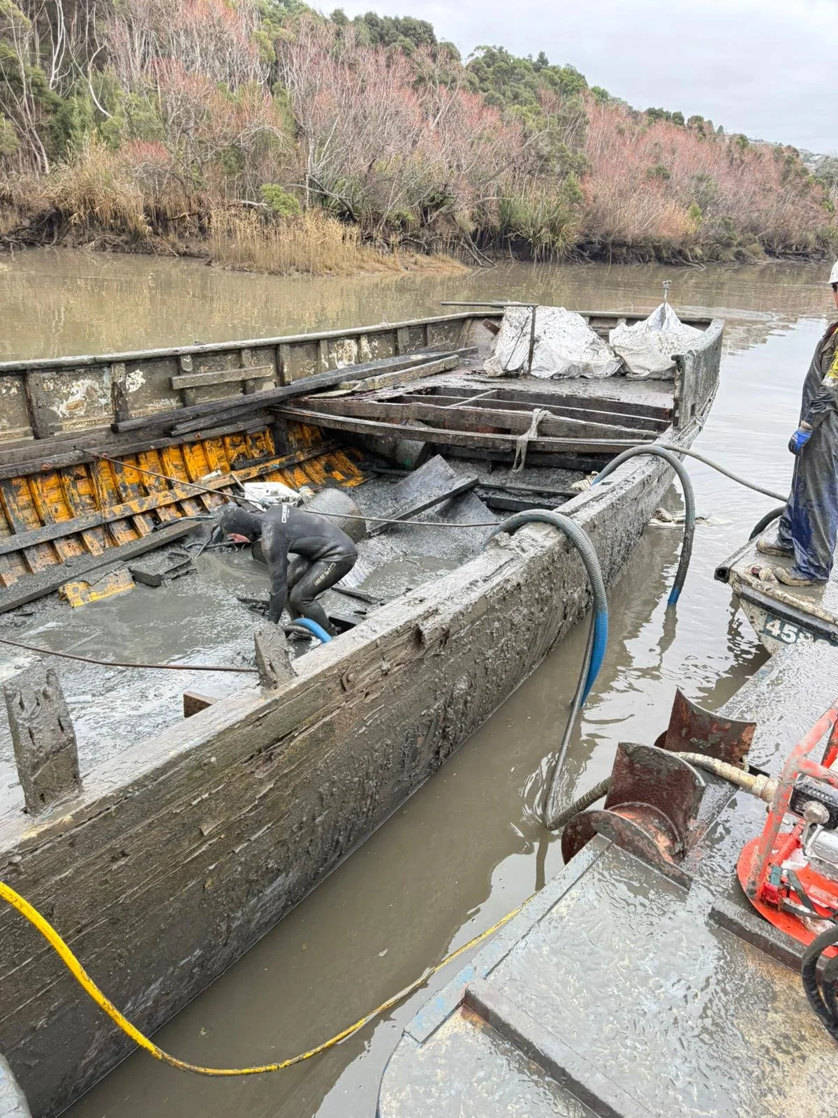 Workers cleaning and repairing a damaged, dilapidated boat on a river, with muddy water and a wooded shoreline in the background.