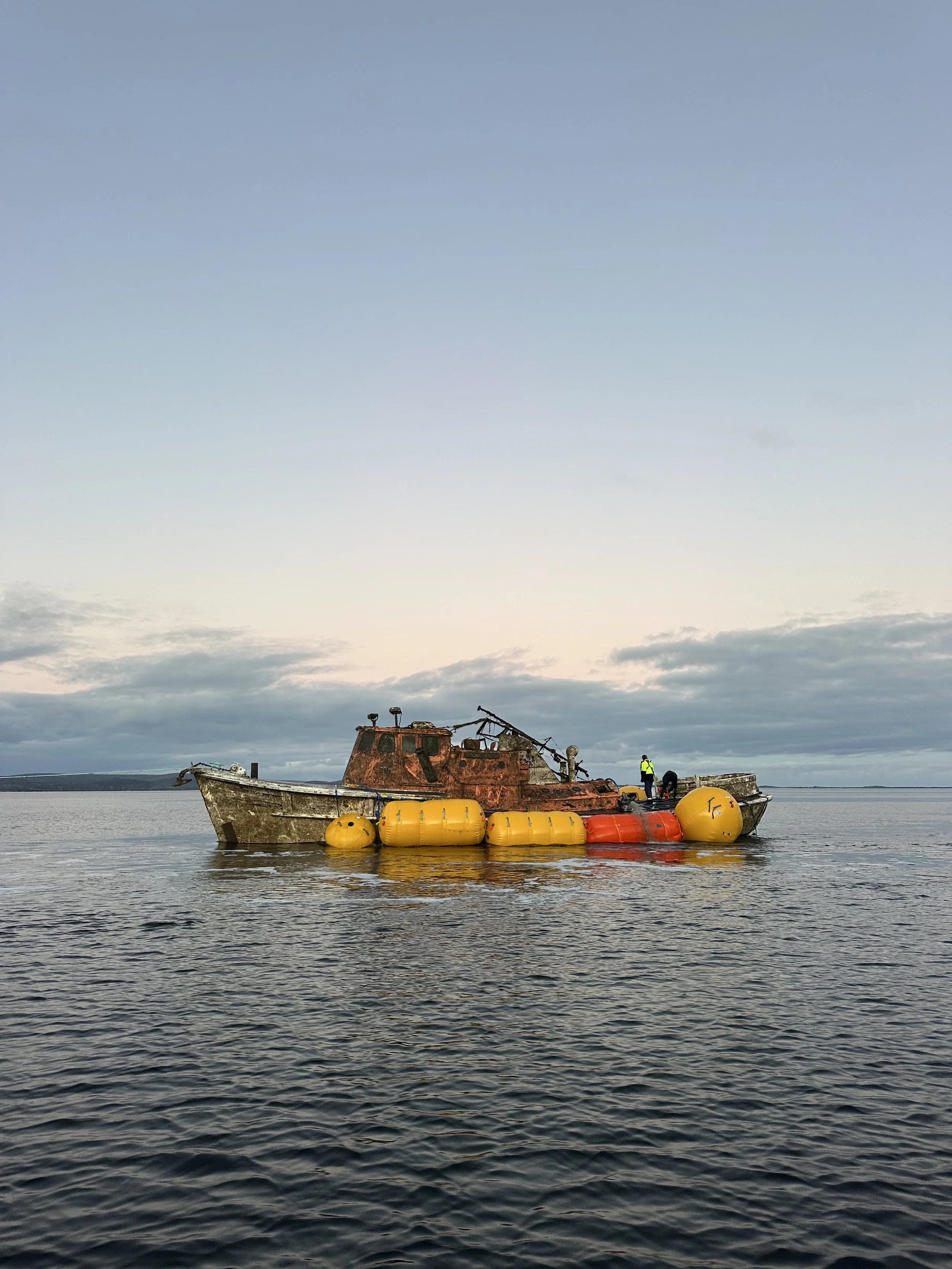 A rusty, abandoned ship surrounded by large yellow and orange buoys on calm water with a cloudy sky in the background.