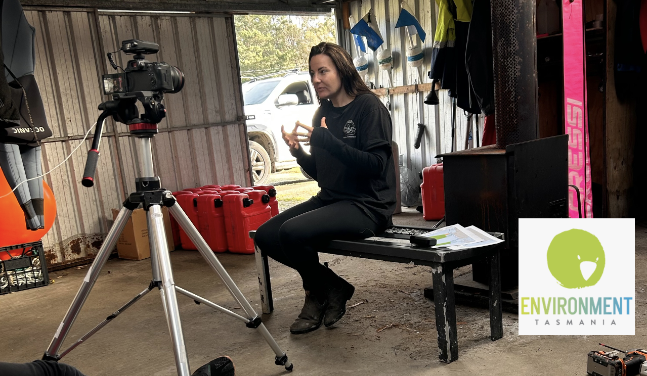 Person speaking on camera in a shed with equipment and gear, filming setup with tripod and camera, Environment Tasmania logo in corner.