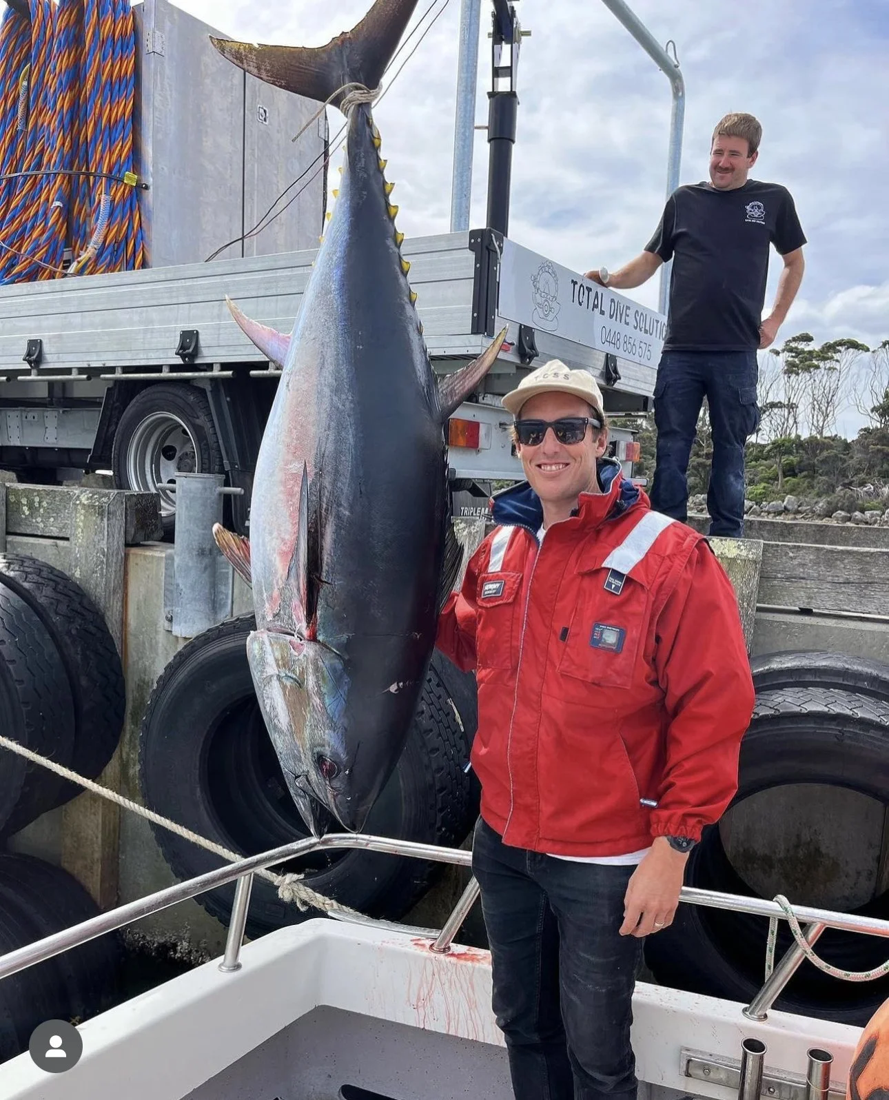 A person in a red jacket standing next to a large tuna fish hanging by its tail on a dock, with another person in the background on a truck.