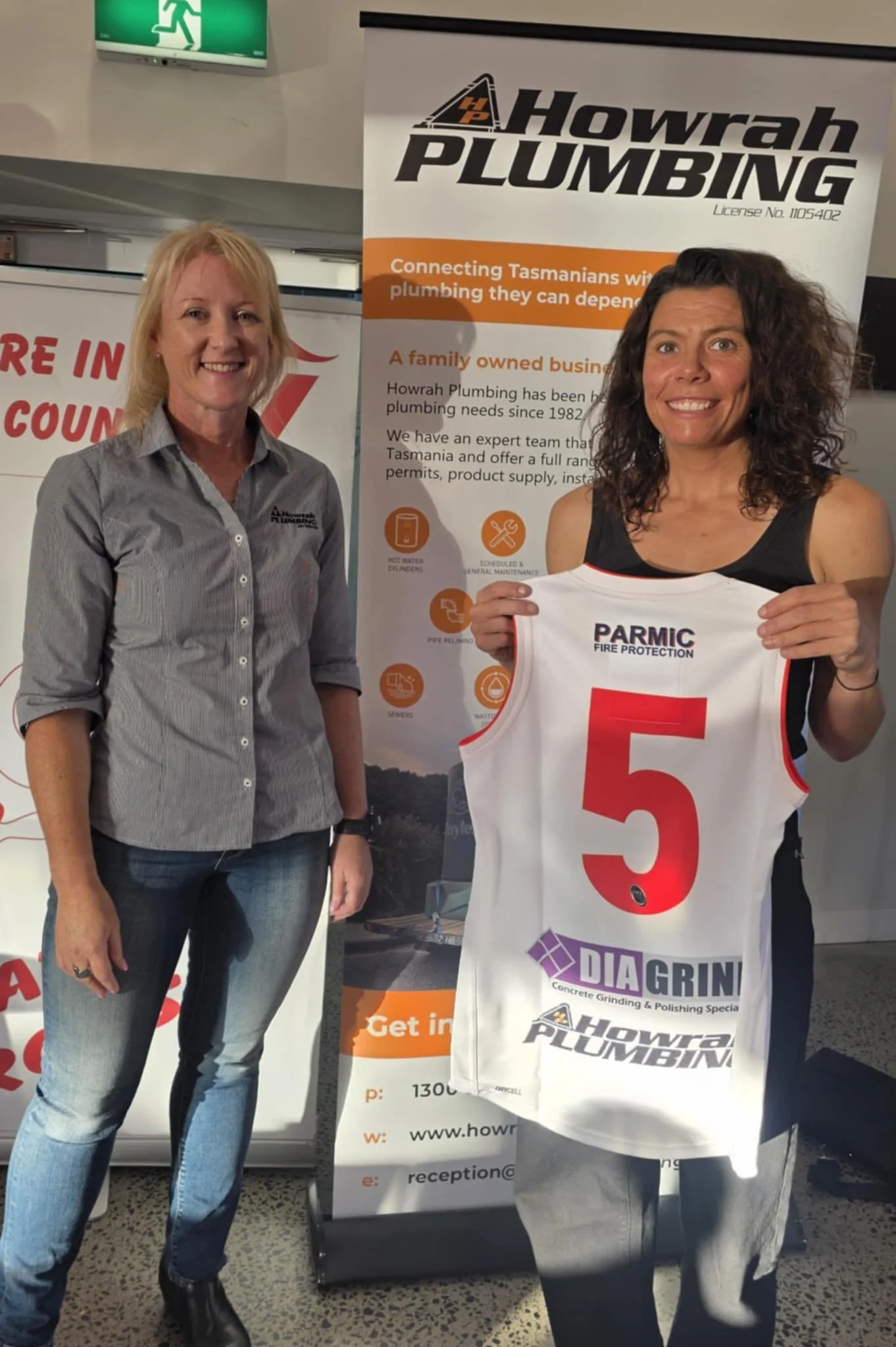 Two women standing in front of a Howrah Plumbing banner. One woman is holding a sports jersey with the number 5 and various sponsor logos, including Parmic Fire Protection and Howrah Plumbing. The setting appears to be indoors with promotional material visible.