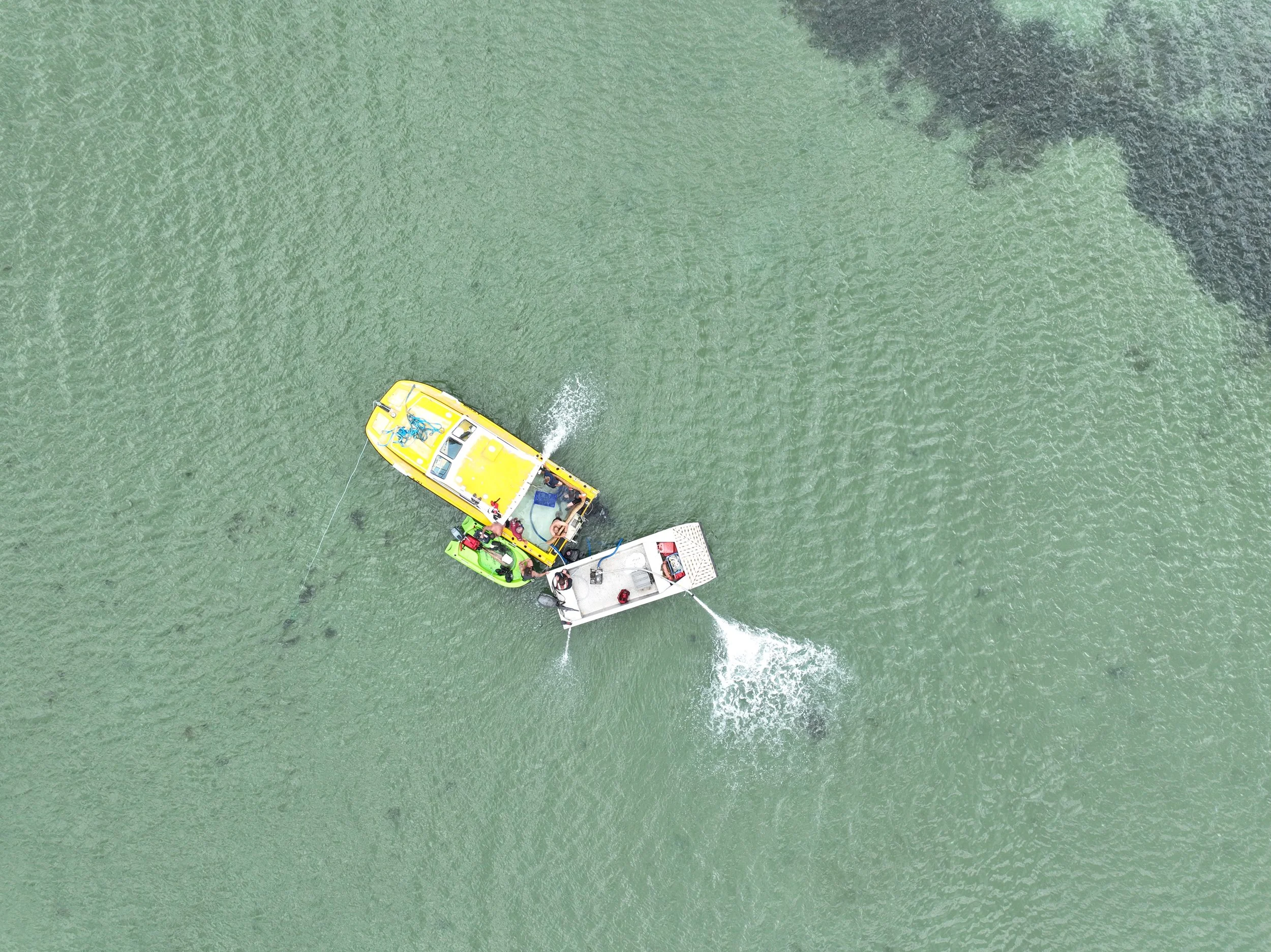 Two boats, one yellow and one white, connected in the water near a rocky shoreline, with a person on the yellow boat and others on the white boat.